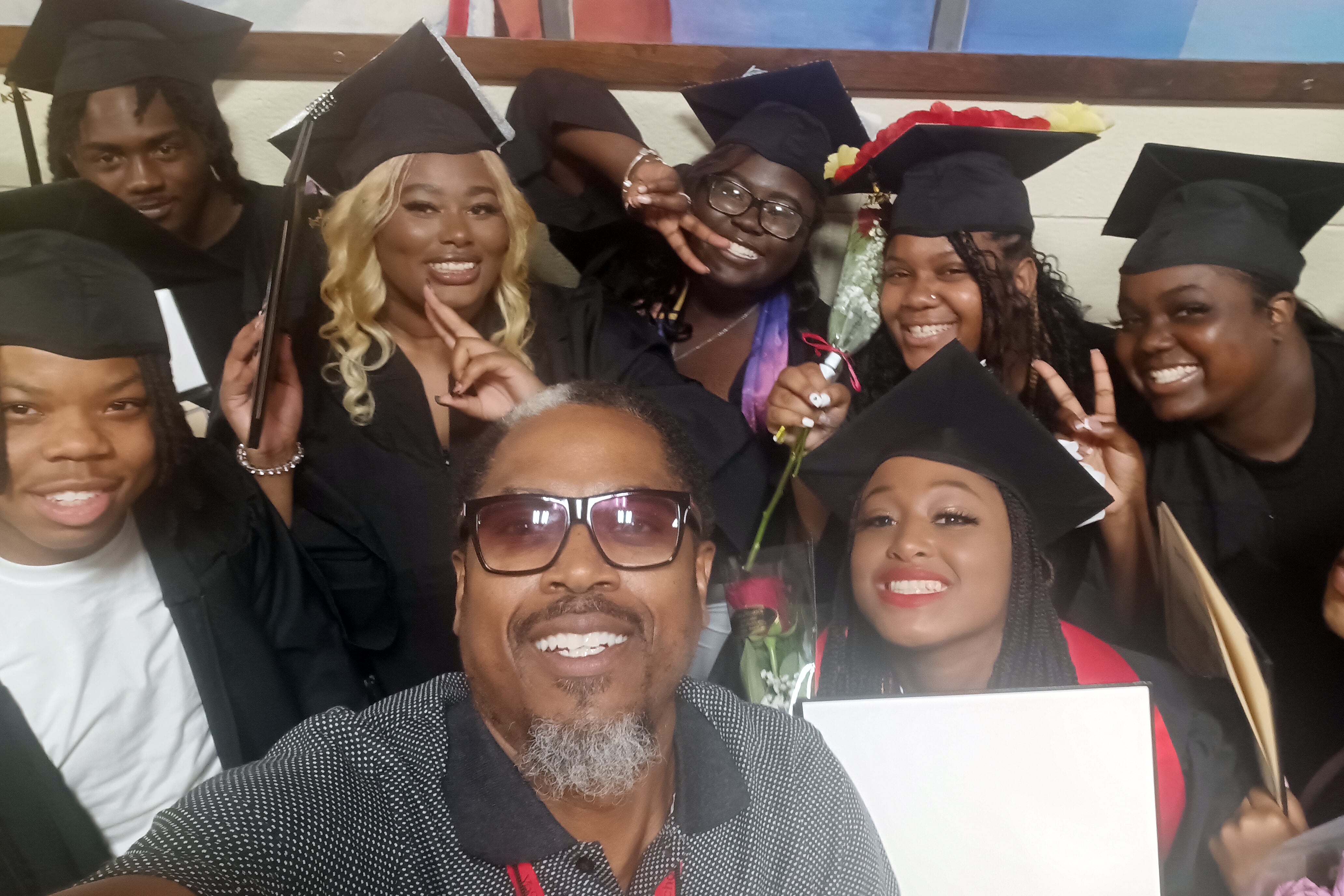 A photograph of a Black man posing with a group of Black high school students all smiling and wearing graduation caps and gowns.