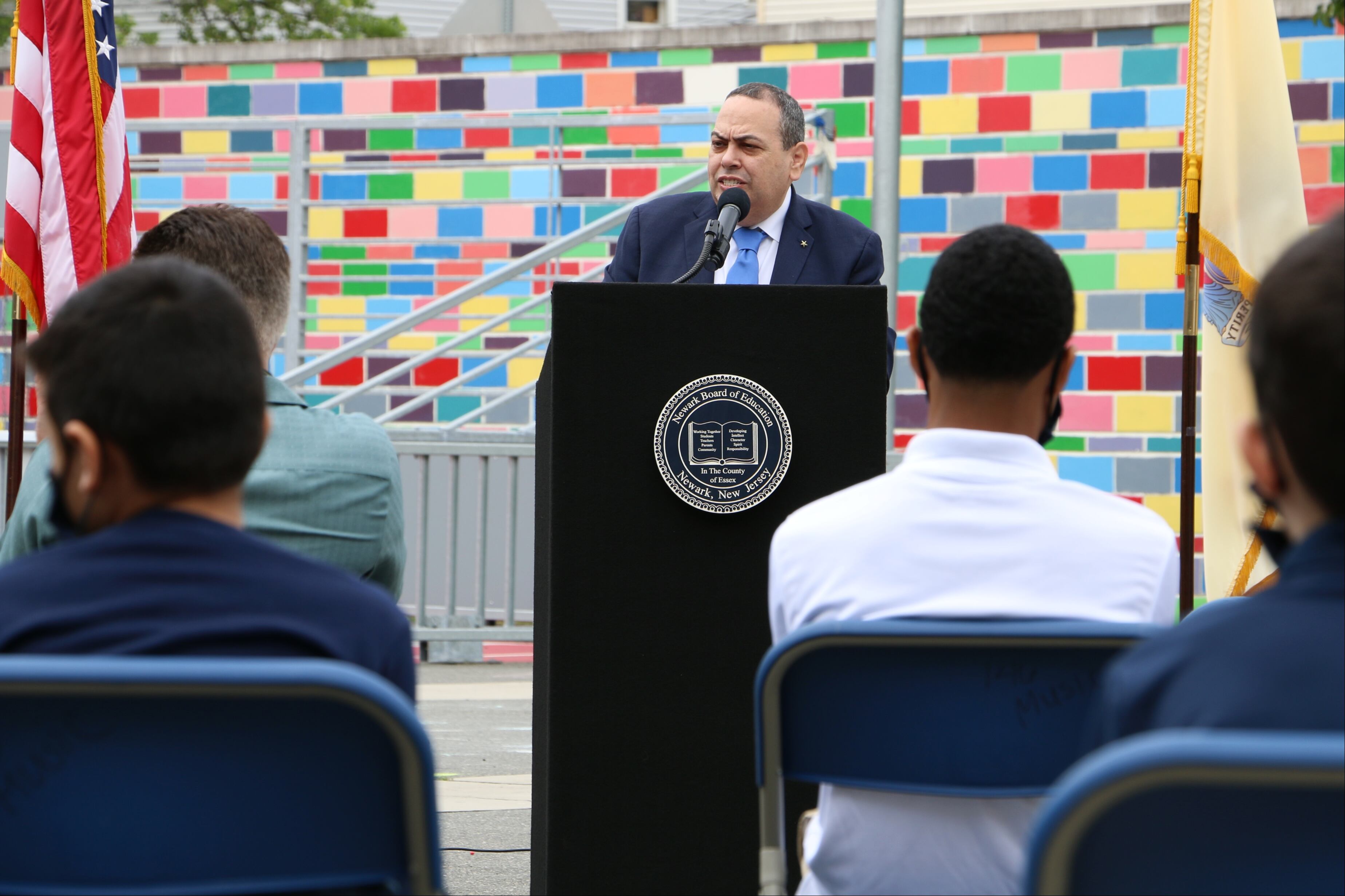 Newark schools Superintendent Roger Leon speaks while standing at a podium against a colorful tile backdrop at First Avenue School, with the backs of several audience members in the foreground and an American flag on the left.