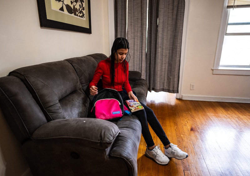A young girl with long dark hair and wearing a red top sits on a couch next to her backpack in a living room.
