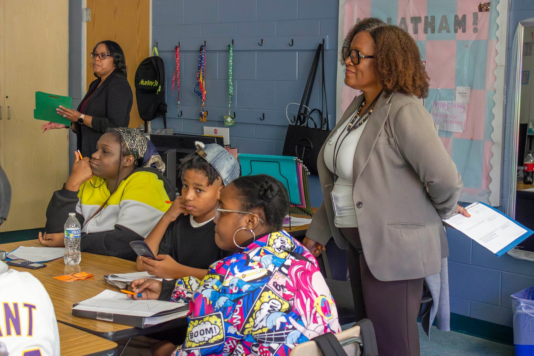 A woman with short brown hair and wearing a grey suit jacket stands next to three students sitting at a wooden desk with another adult in the background.