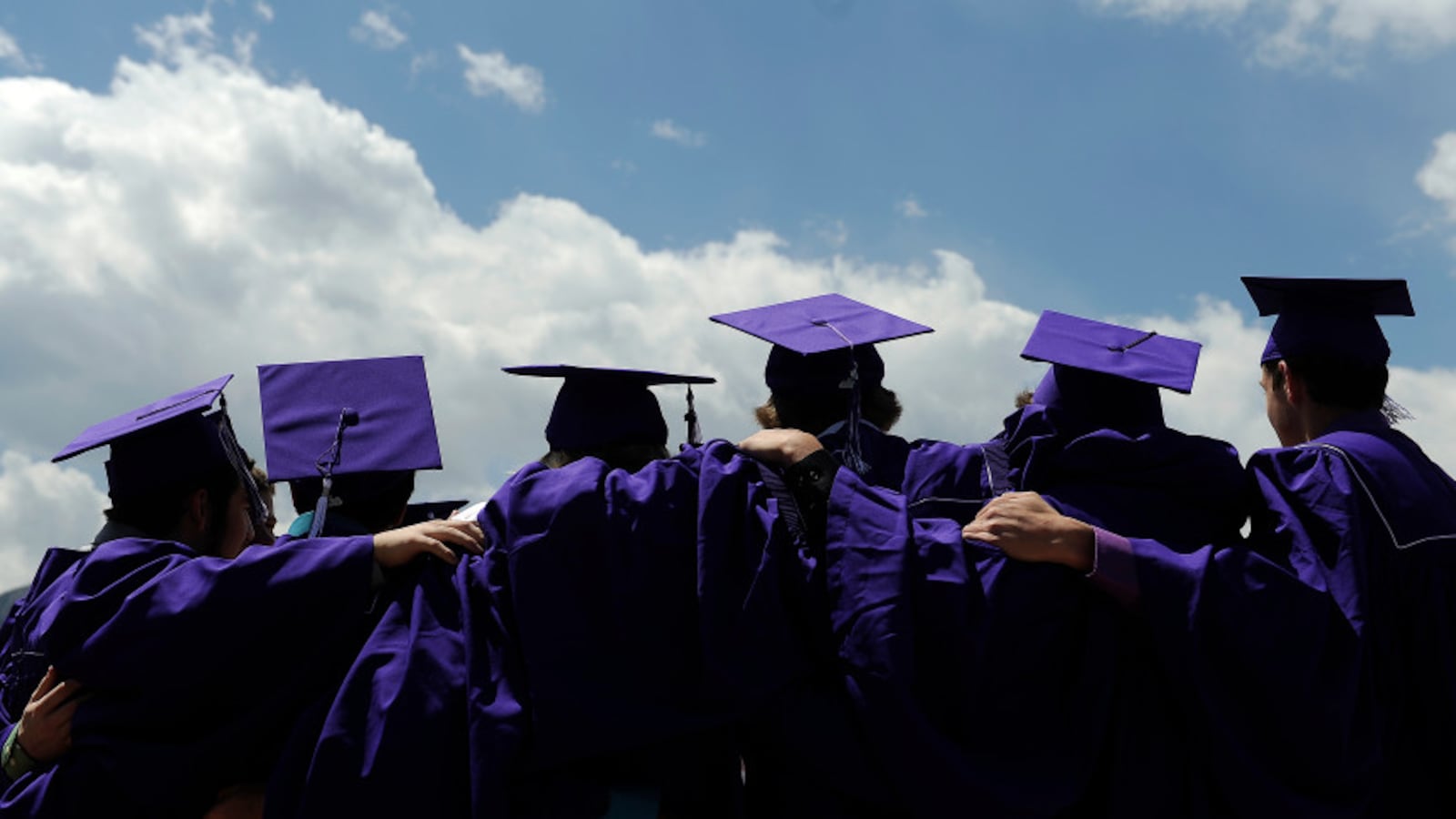 The backs of six students in purple caps and gowns in a line with their arms around each others’ shoulders.