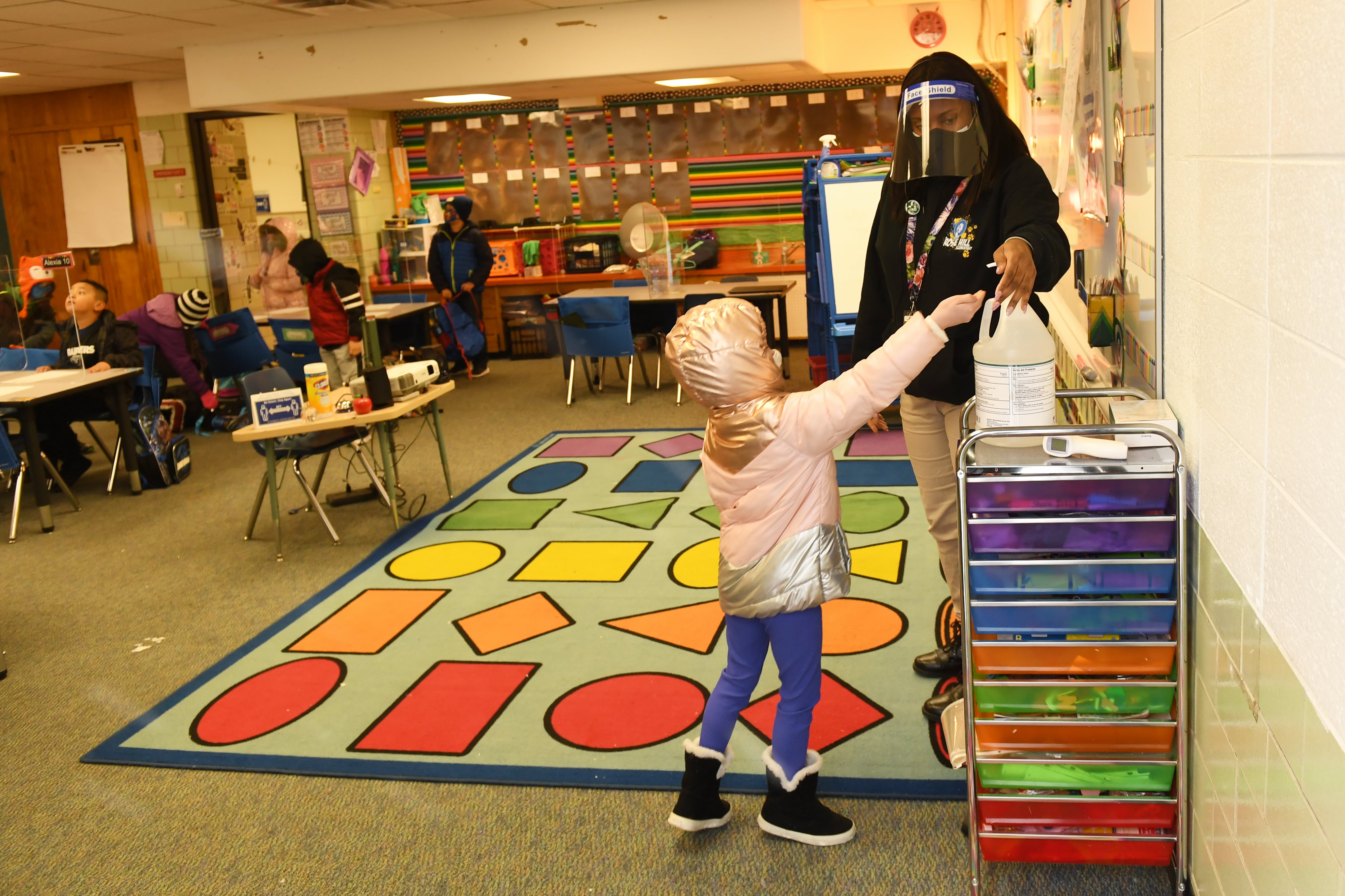 A teacher helps a young student get a squirt of hand sanitizer in a colorful elementary classroom.