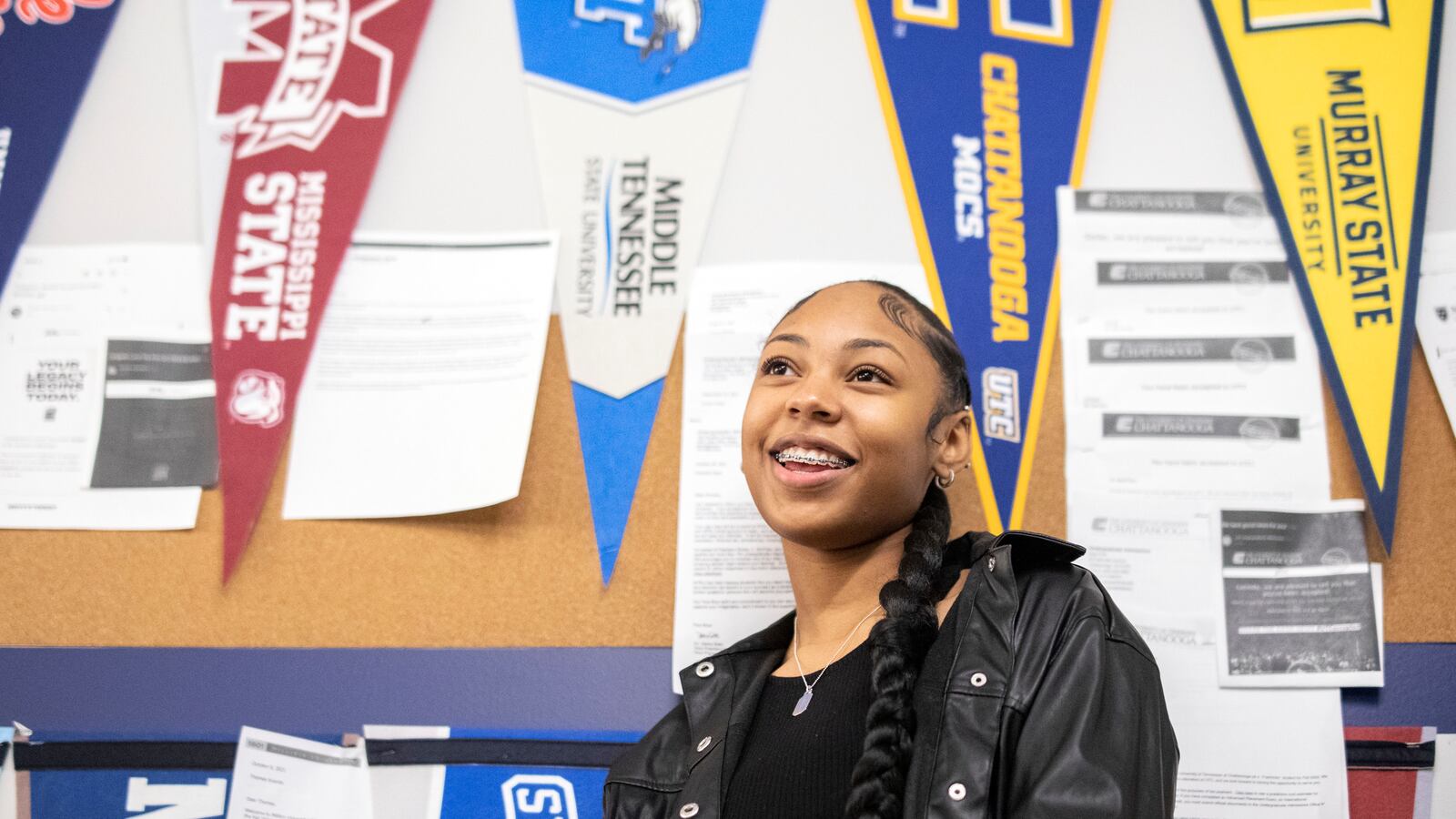 A young woman, wearing a black jacket and shirt and showing rows of braces on her teeth, smiles in front of a classroom wall adorned with college banners.