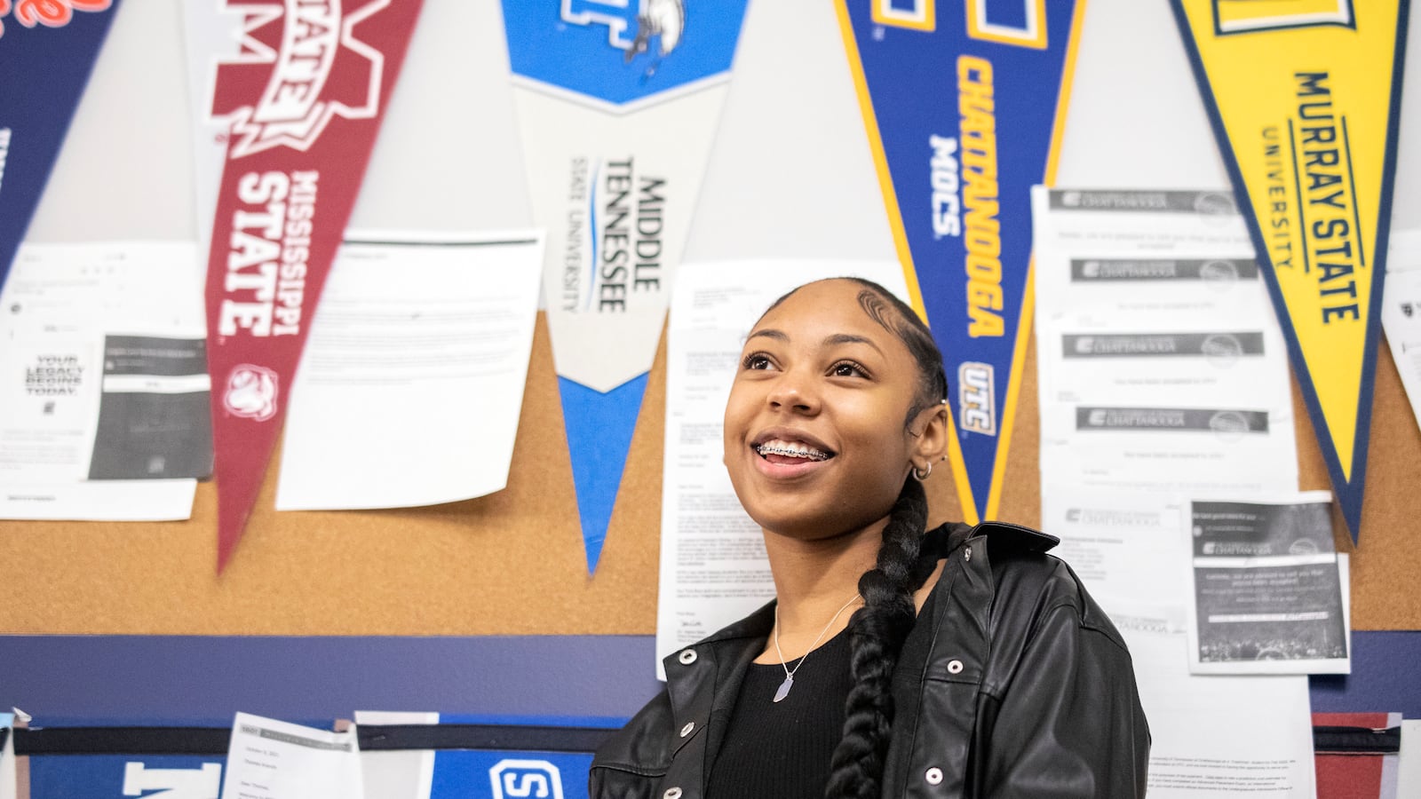 A young woman, wearing a black jacket and shirt and showing rows of braces on her teeth, smiles in front of a classroom wall adorned with college banners.