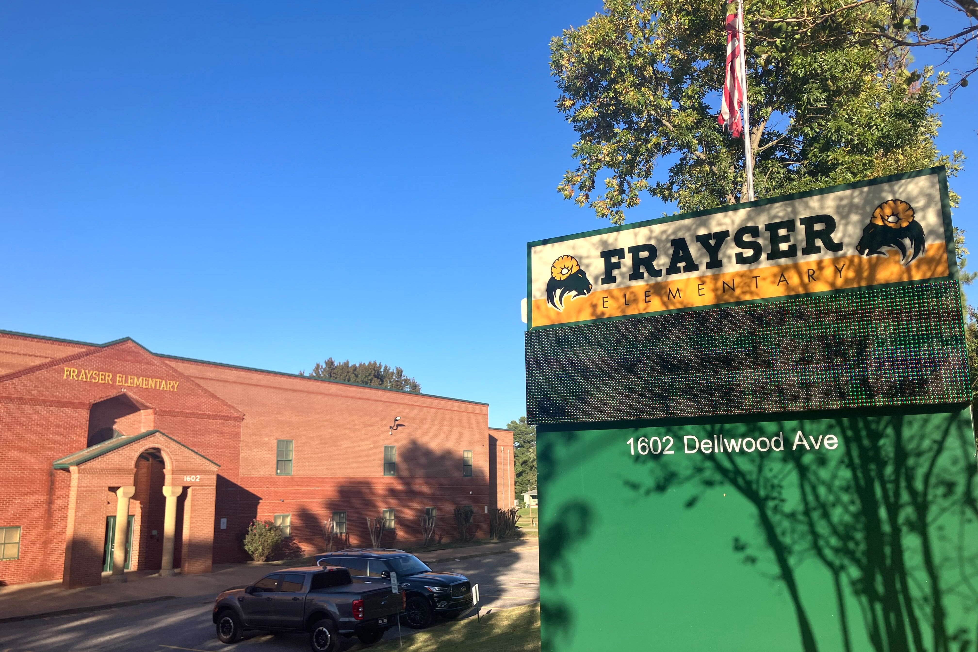 A photograph of a large green and black sign in front of a brick school building with two cars parked in the parking lot and a clear blue sky in the background.