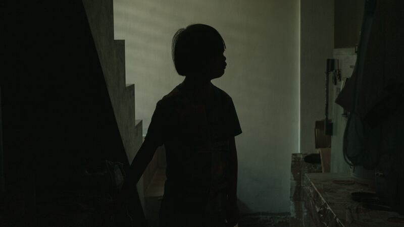 A boy stands silhouetted in a dark hallway next to a counter and a set of stairs.