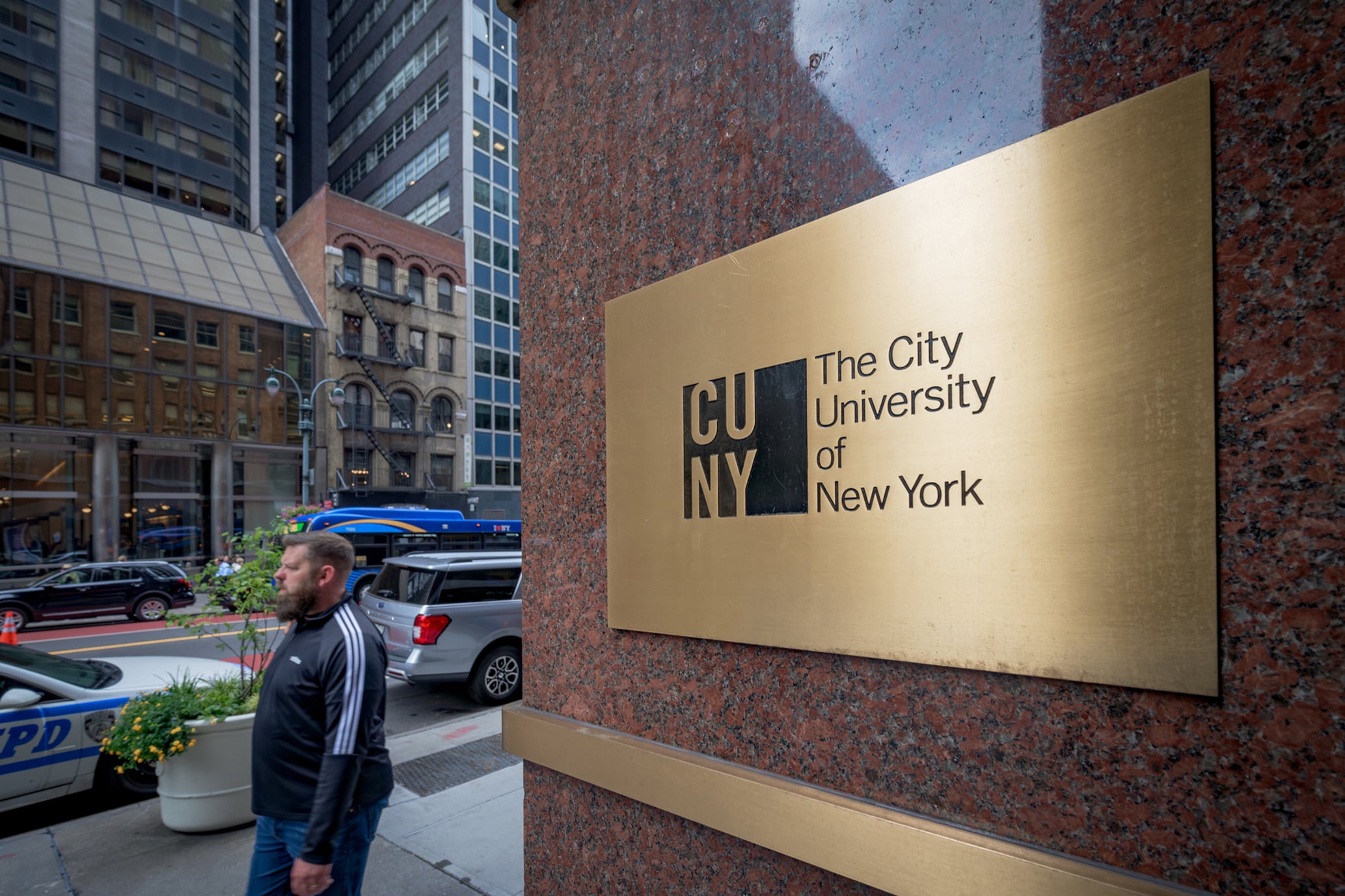 A person in a track suit walks by a gold and black plaque at the entrance of CUNY Central with cars along the street in the background.