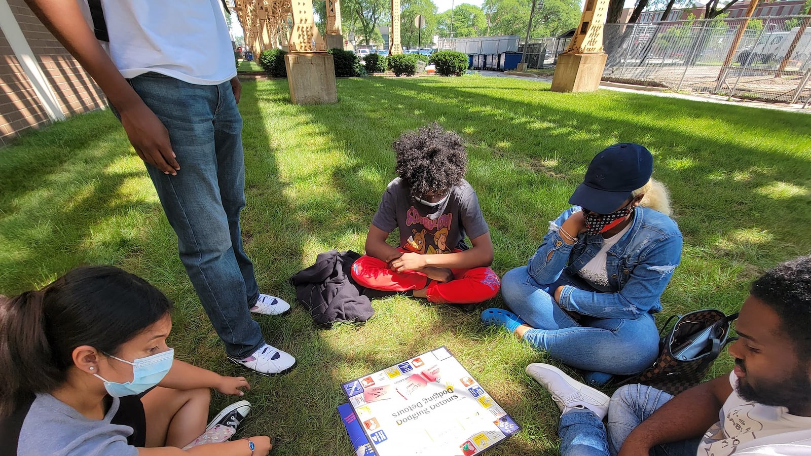 Students sit and play a board game outdoors, each wearing protective masks