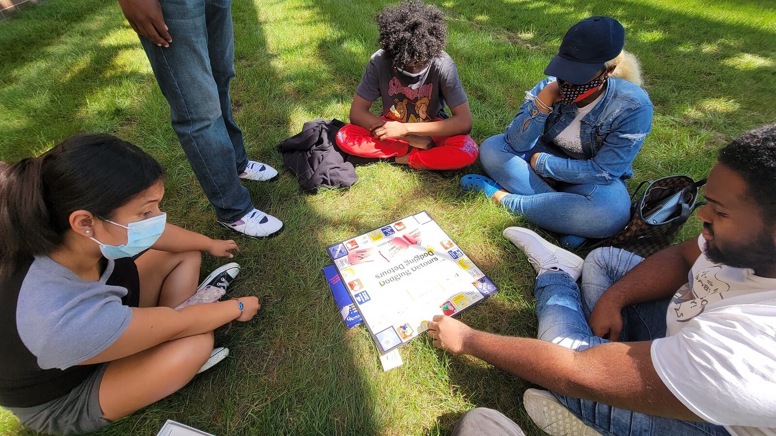 Students sit and play a board game outdoors, each wearing protective masks