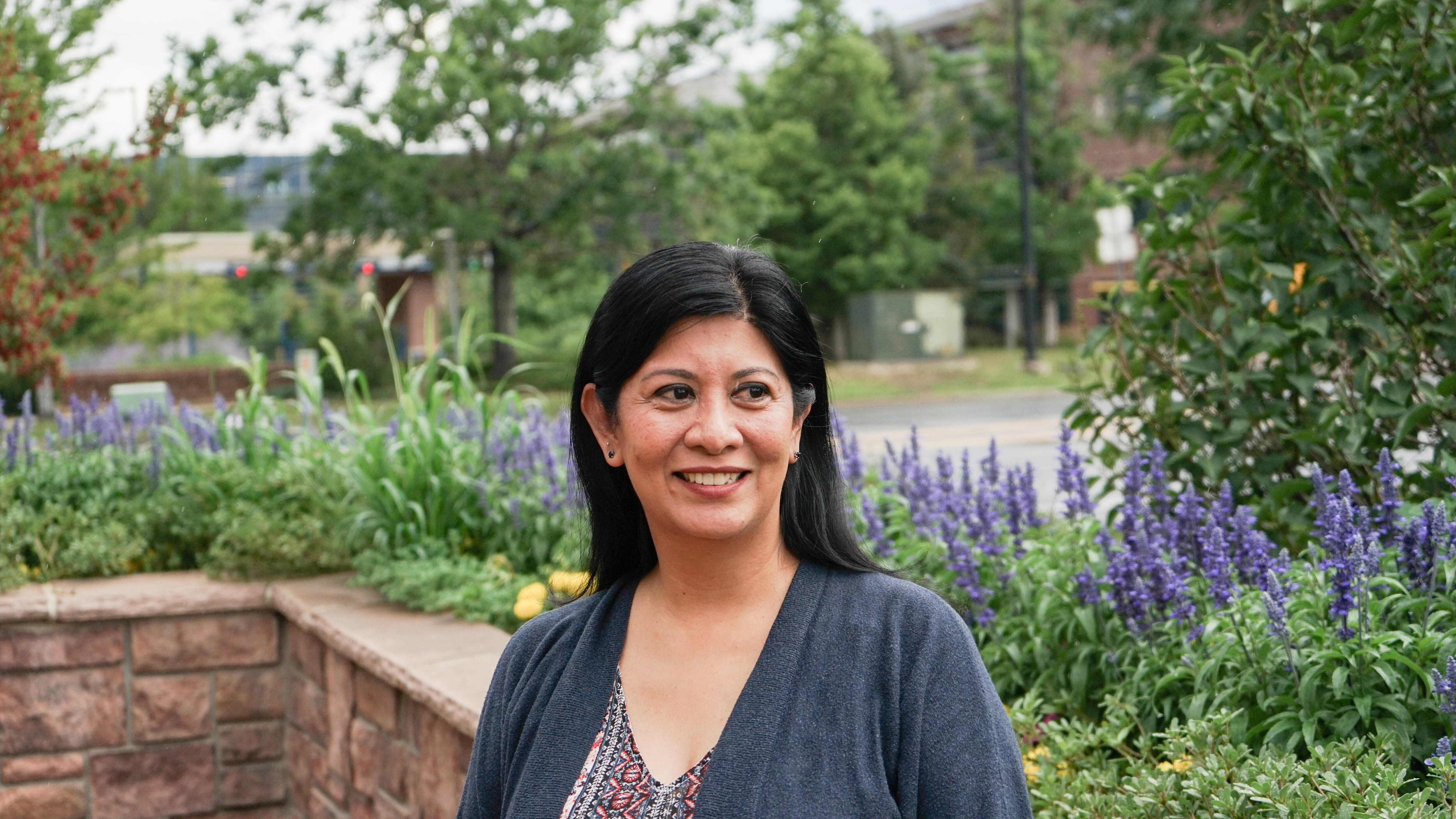 A woman with shoulder-length dark hair poses for a portrait outside a building. She’s in a park-like setting with lots of purple flowering plants around her.