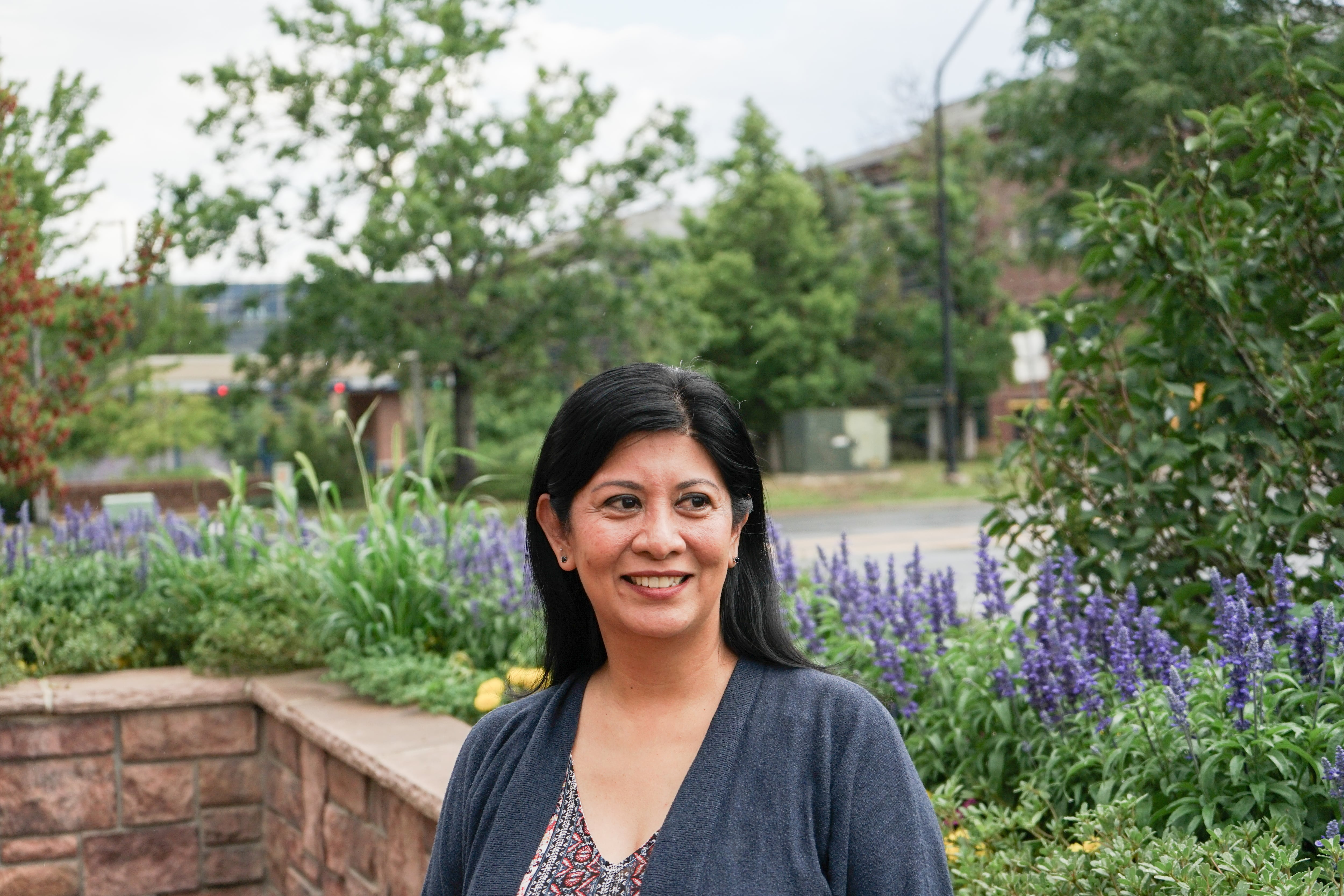 A woman with shoulder-length dark hair poses for a portrait outside a building. She’s in a park-like setting with lots of purple flowering plants around her.