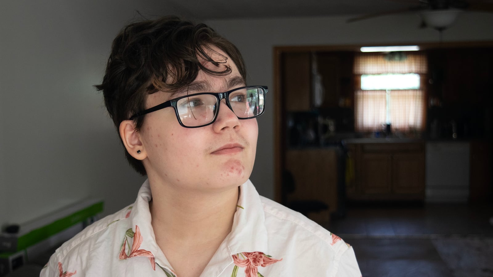 A portrait of a youth in glasses and wearing a white shirt with pink flowers. In the background, a kitchen is dimly visible.