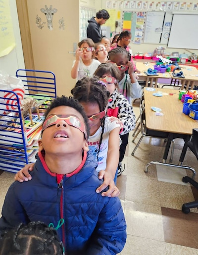 A line of young students in a classroom with an adult in the background.