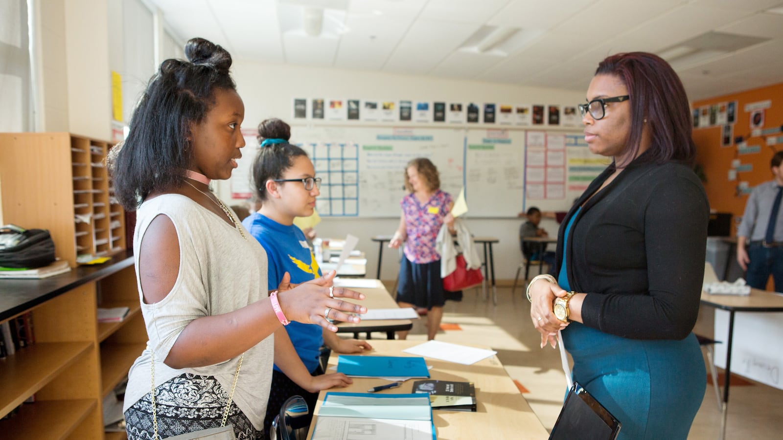 A student wearing a white shirt talks to a teacher wearing a black shirt inside of a classroom.