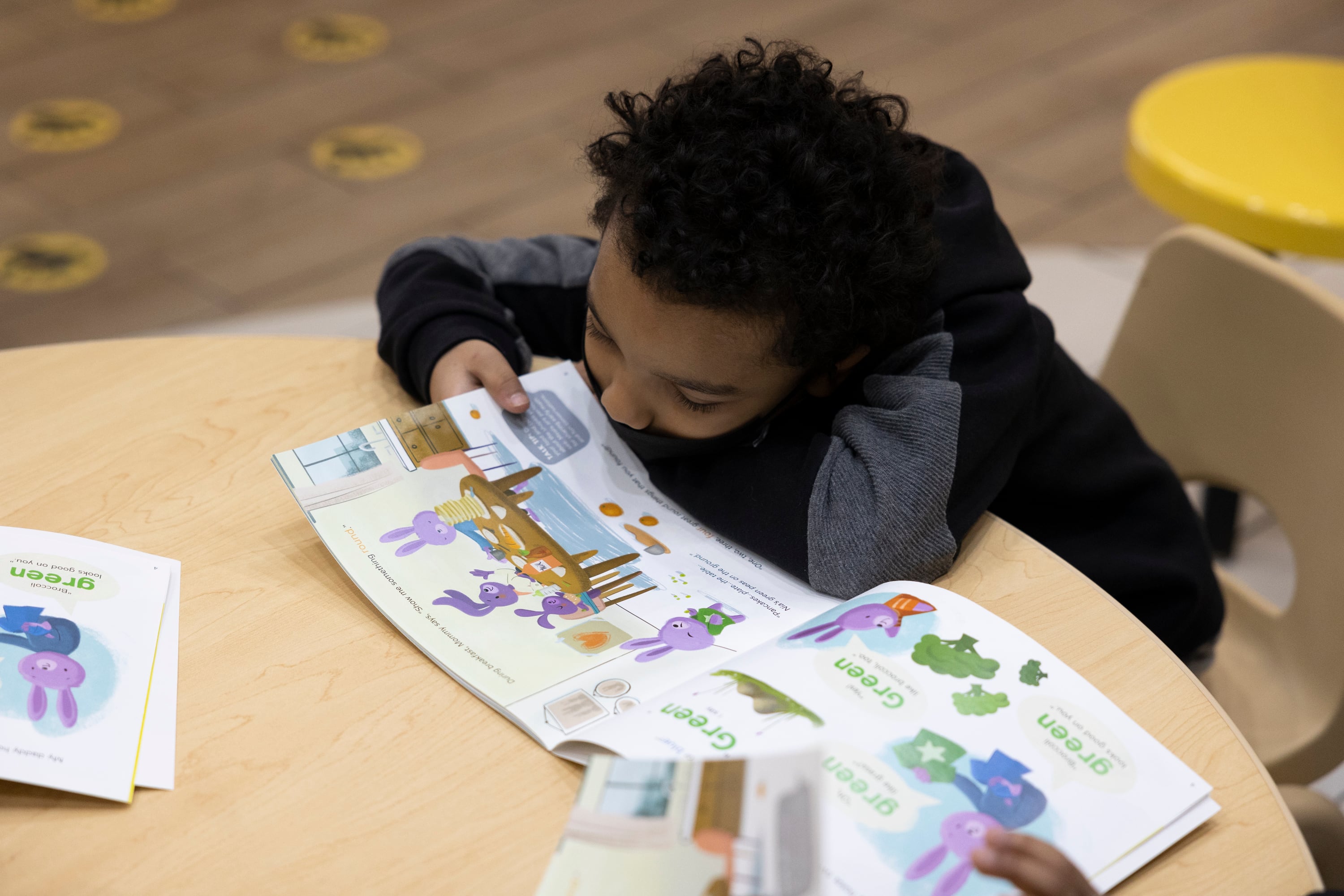 A little boy reads a children's book at a small wooden table.