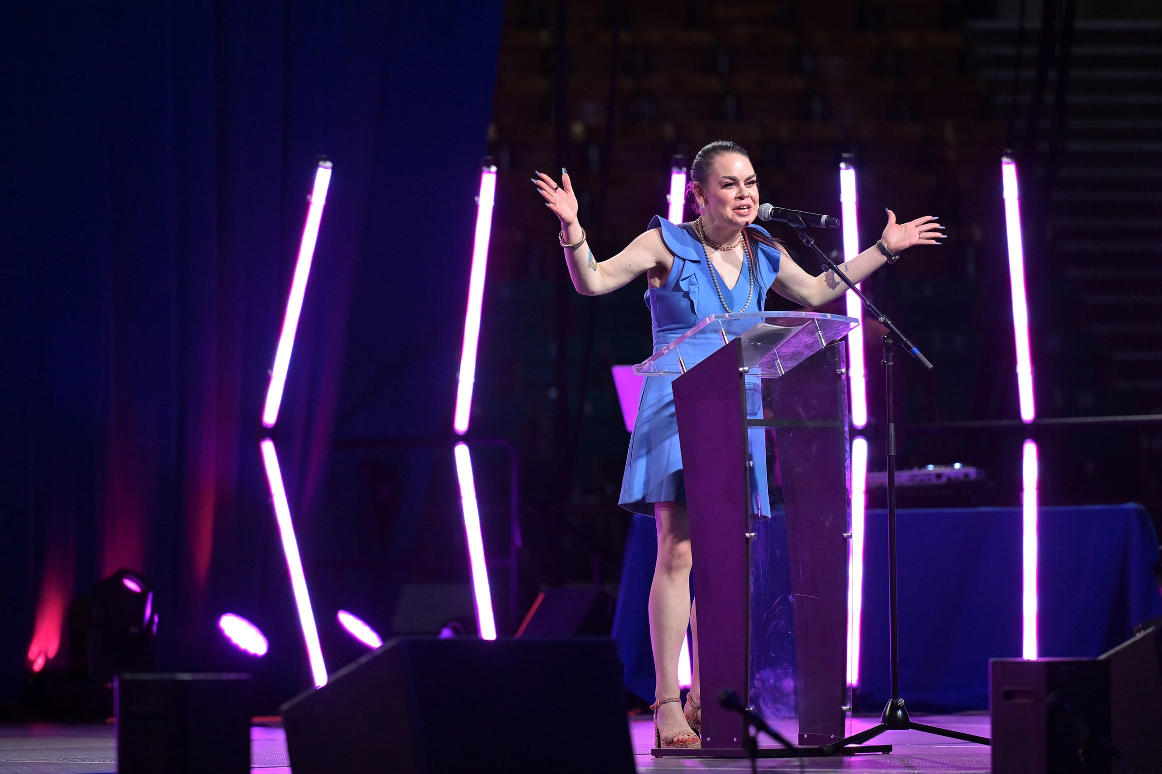 A photograph of a woman speaking from behind a podium on stage with purple lights in the background.