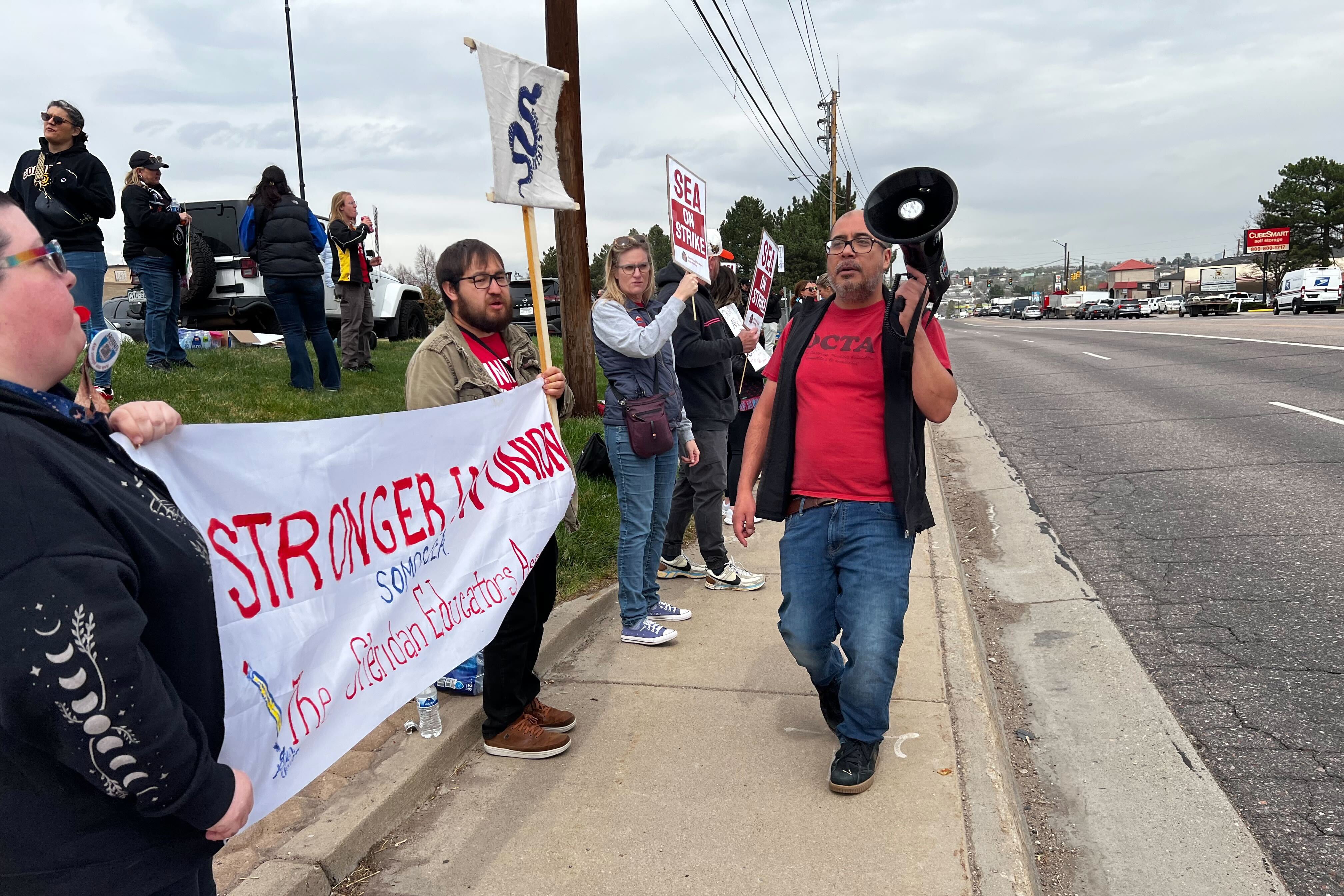 A group of adults stand outside some are holding signs or banners or a megaphone.