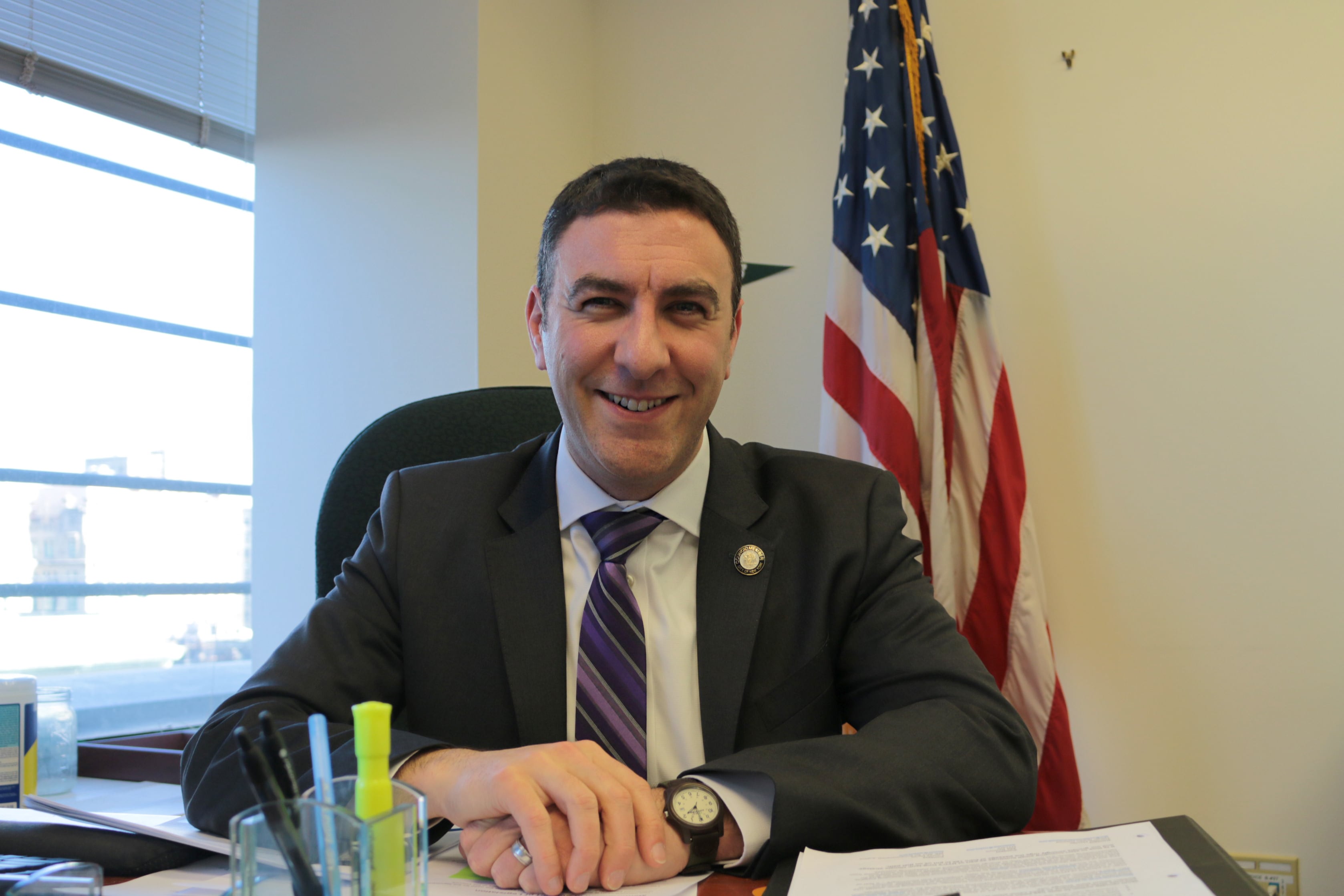 A photograph of a man in a suit sitting at a desk smiling at the camera with an American flag in the background.