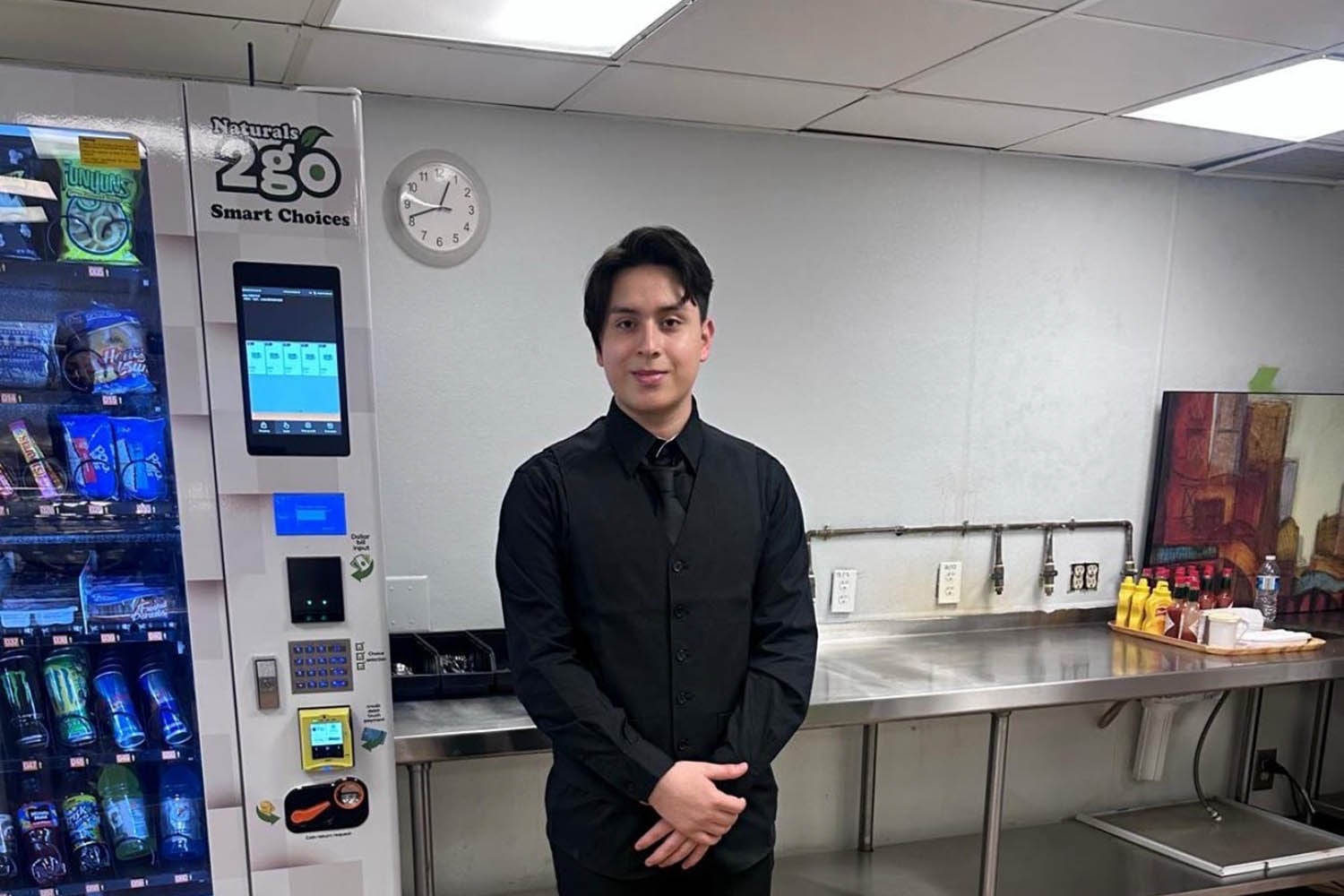 A high school student in an all black suit poses for a portrait in a cafeteria.
