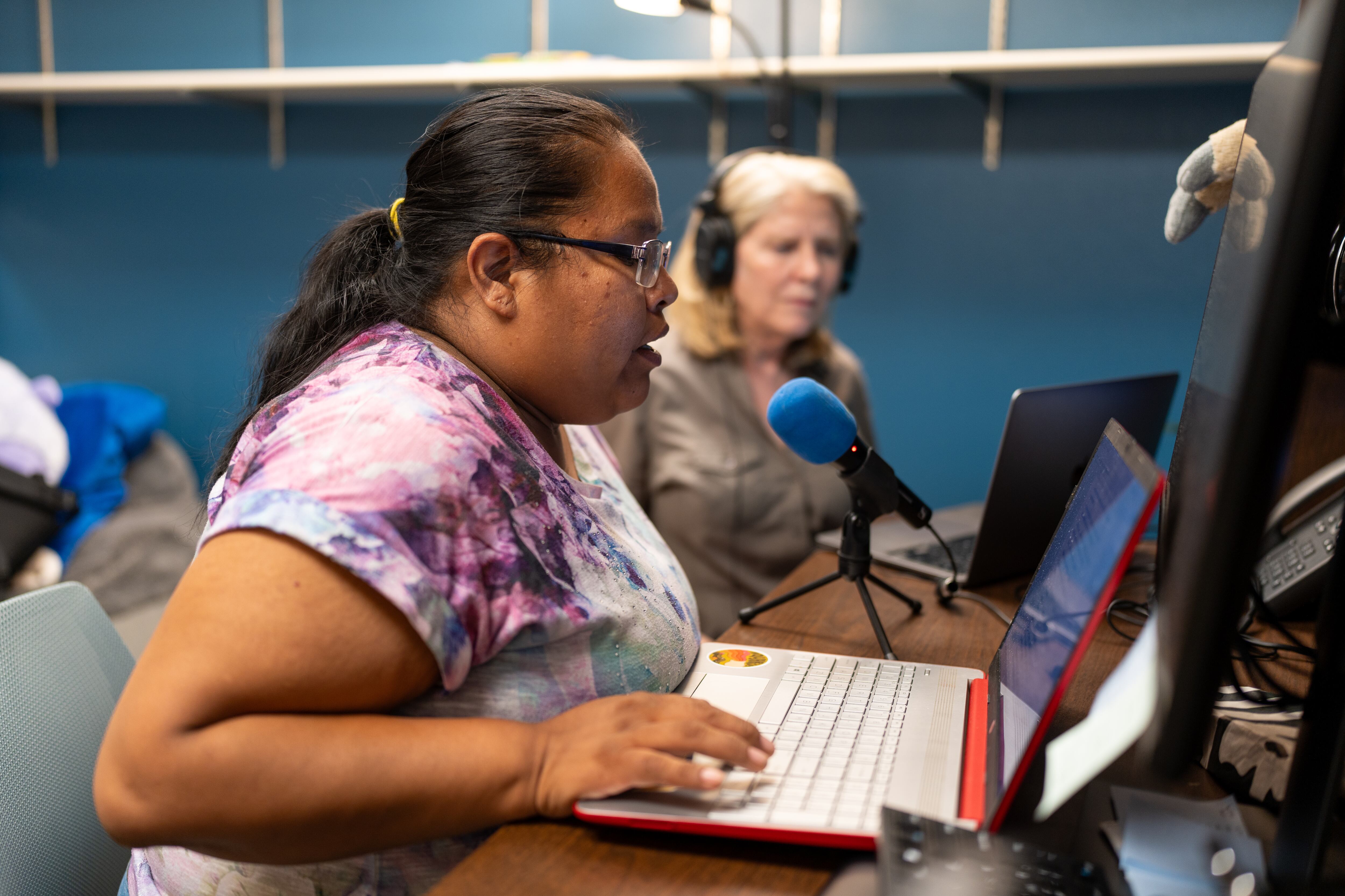 A woman with black hair and glasses in a purple, white, and blue tie dye shirt talks into a microphone.