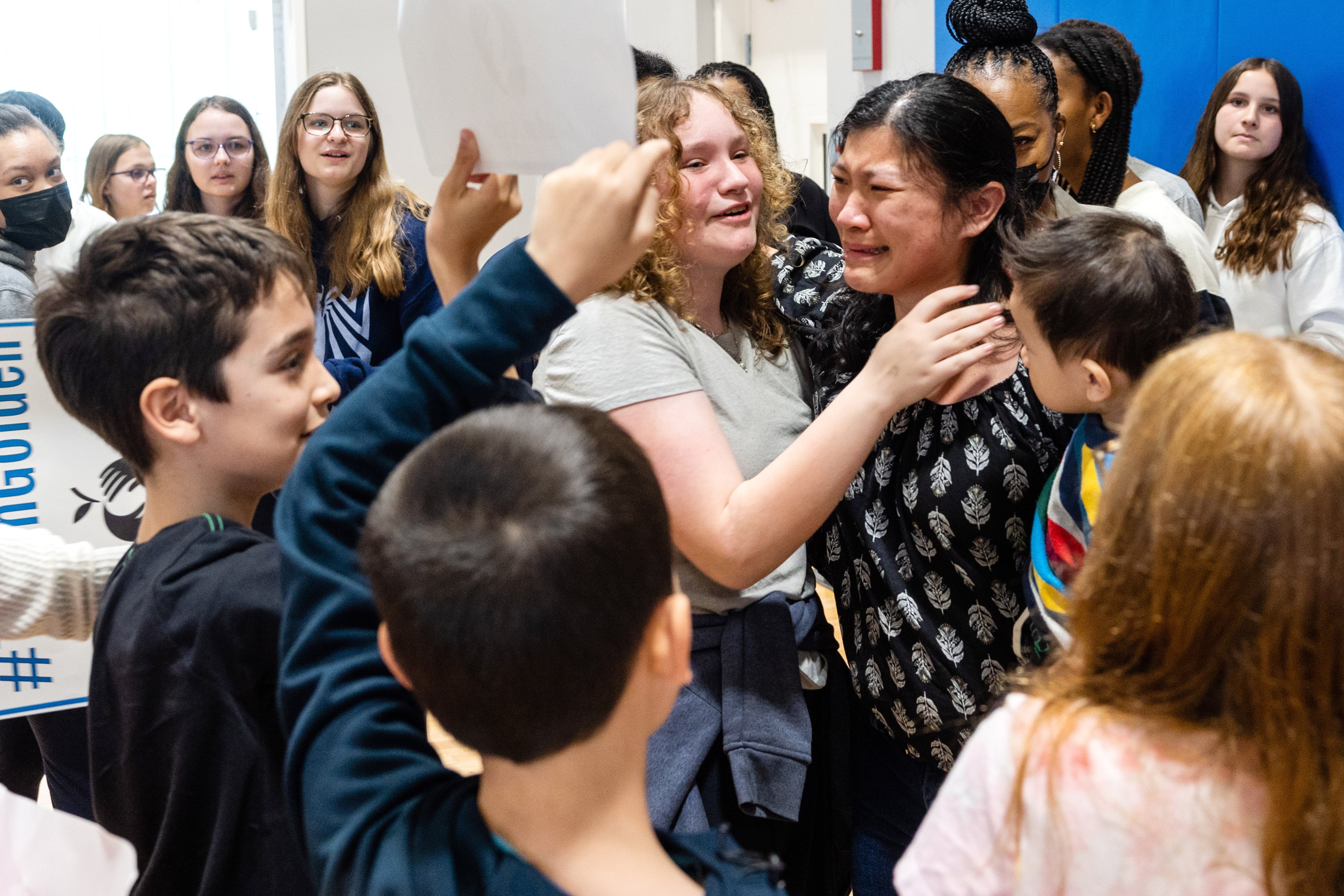 A woman in a black top cries and is hugged and surrounded by students