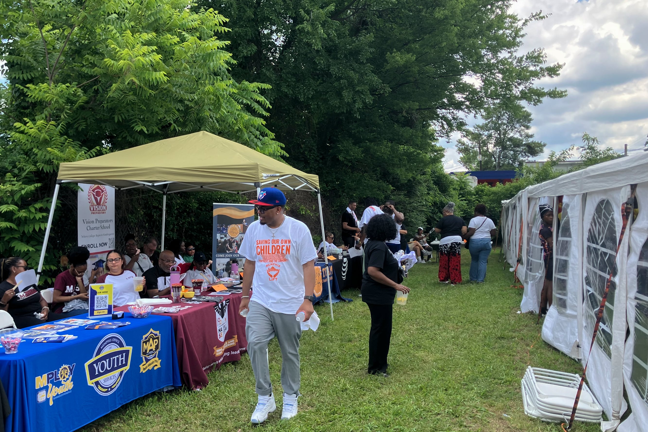 A photograph of a Black male wearing a white shirt that reads "Saving Our Own Children" walks by multiple stands on a lawn.