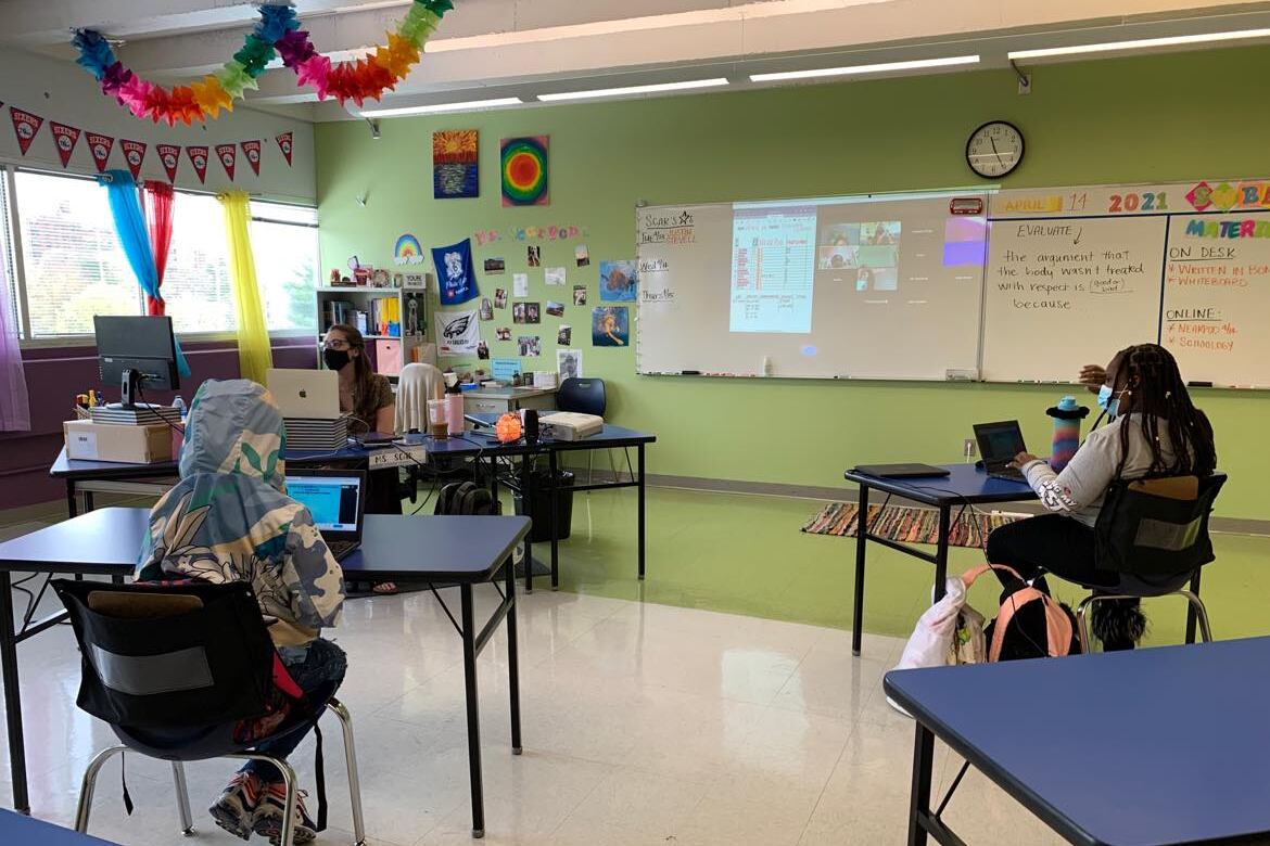 Students sit at large tables, spaced apart in a classroom.