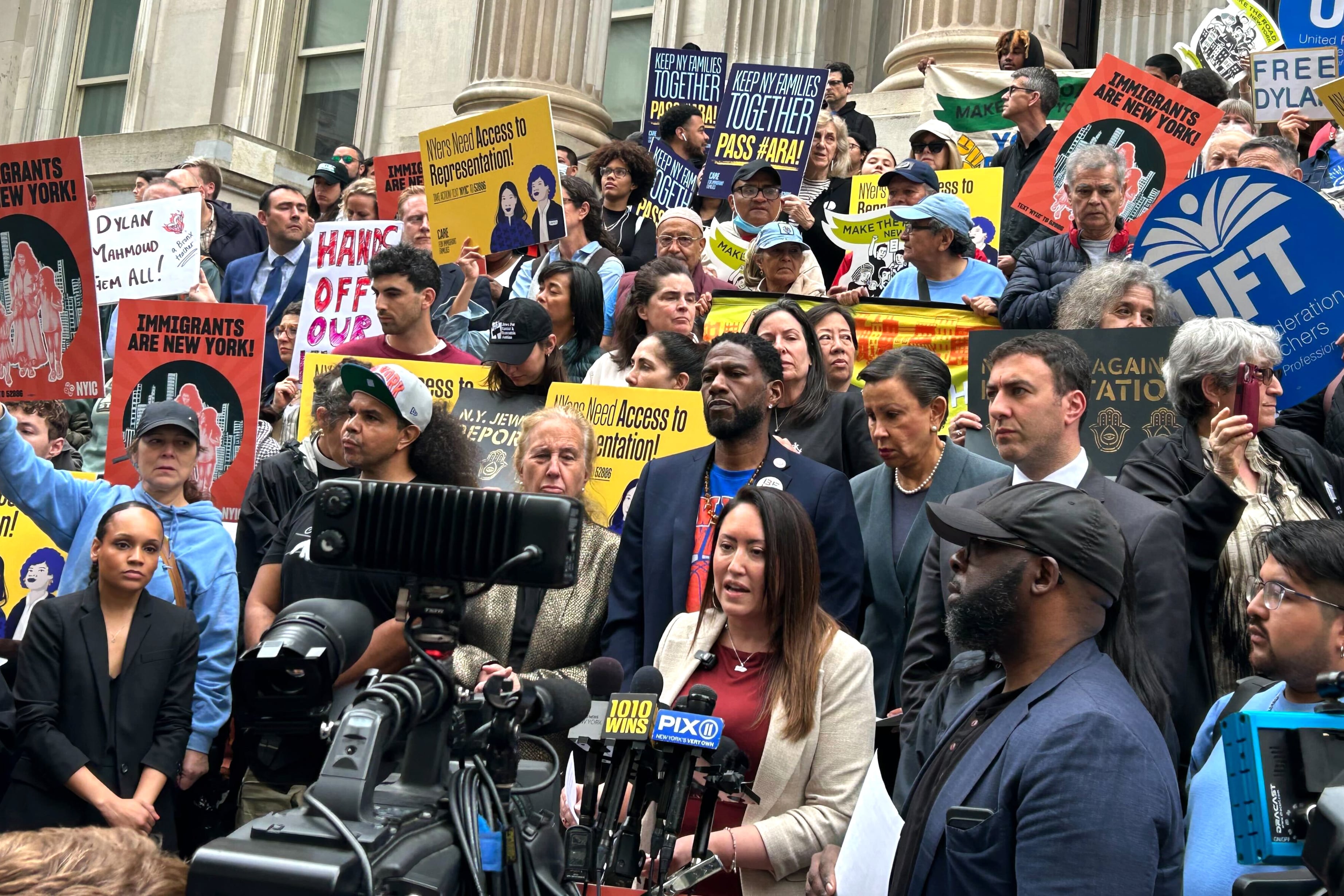 A woman in a red top and light jacket speaks with lots of people, some holding signs, standing behind her.