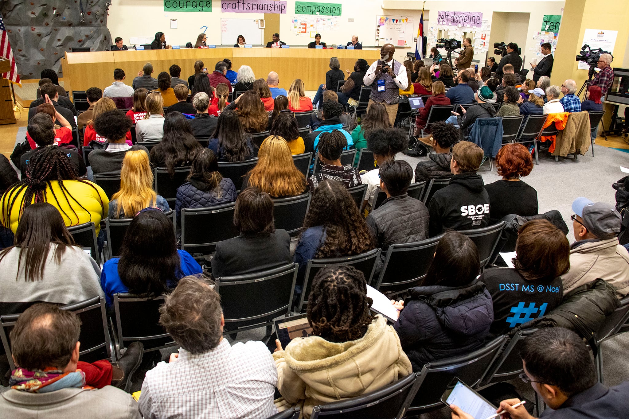 People seated in folding chairs listen to a school board meeting in a gymnasium.