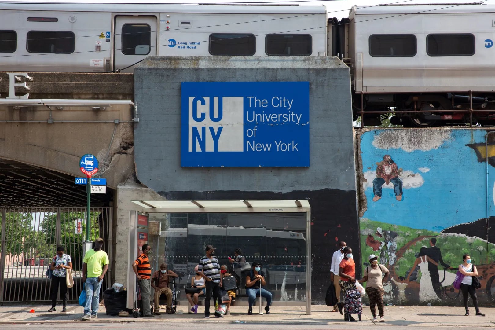A sign for a CUNY campus with NYC silver subway trains running above ground over it.
