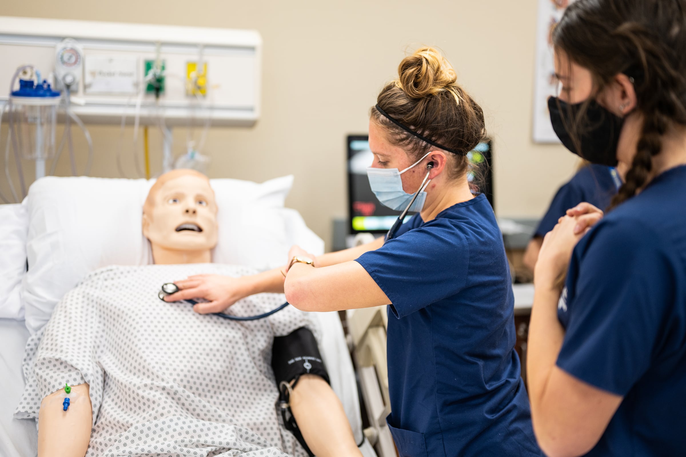 Two nursing students dressed in blue scrubs and with face masks use a black stethoscope on a dummy.