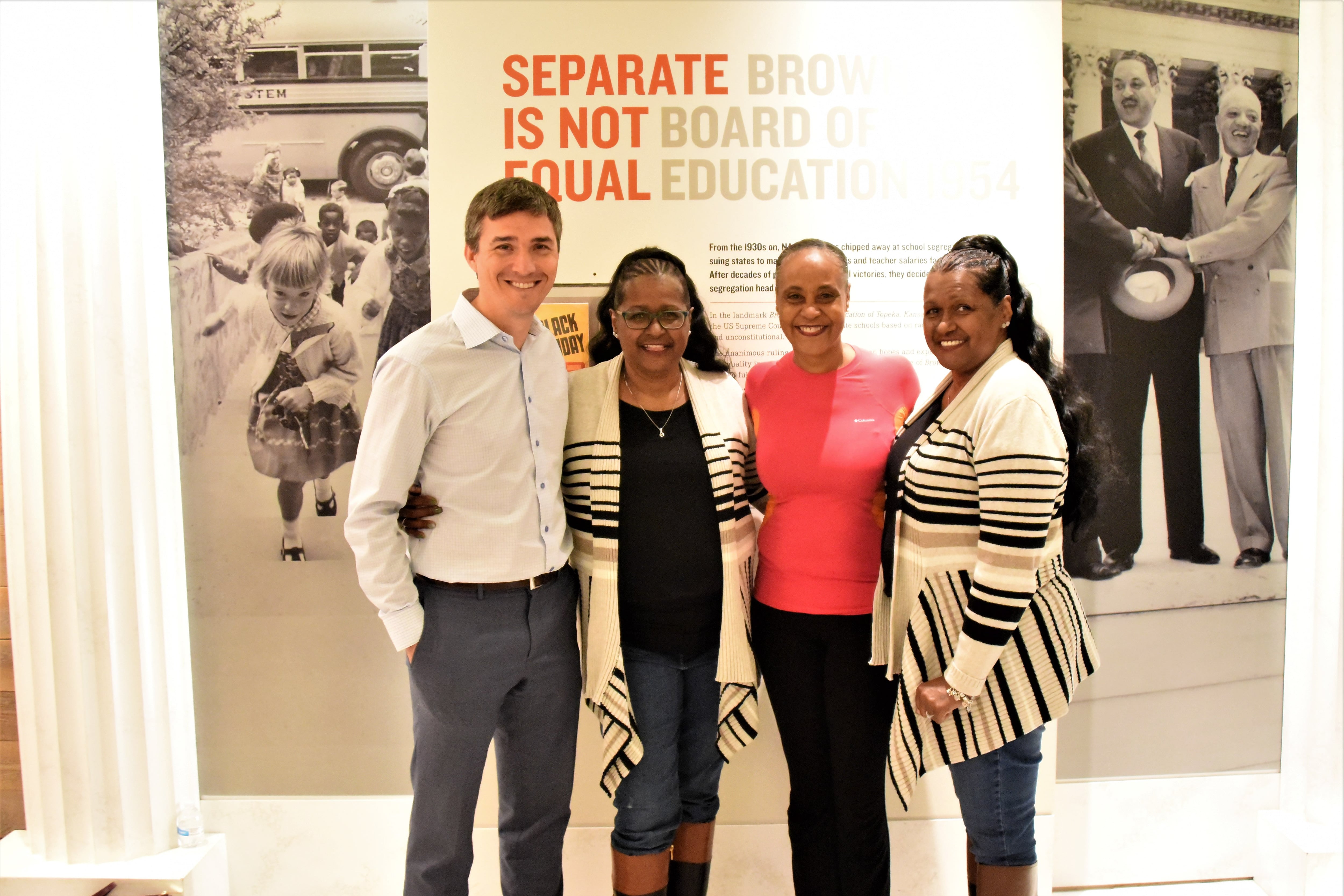 Four people stand together posing for a photograph in front of an exhibition panel in a room.