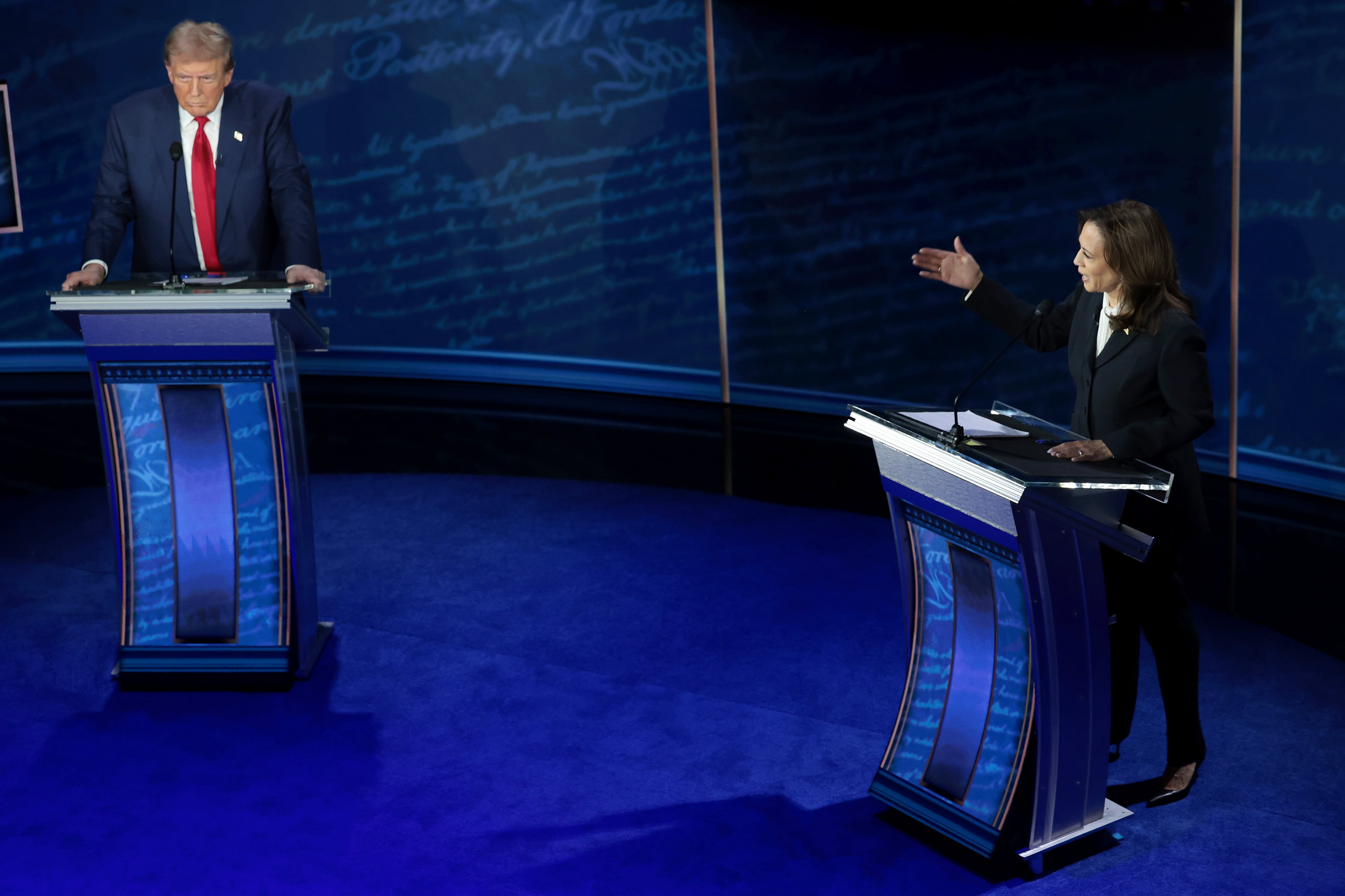 Two adults in suits stand behind blue podiums in a blue room.