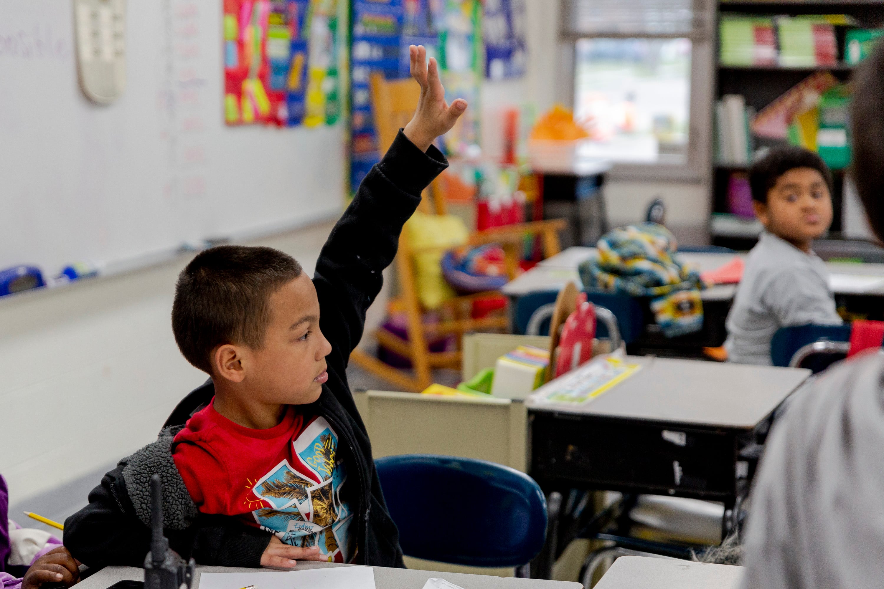 a student raises his hand intently sitting in front of a desk full of papers