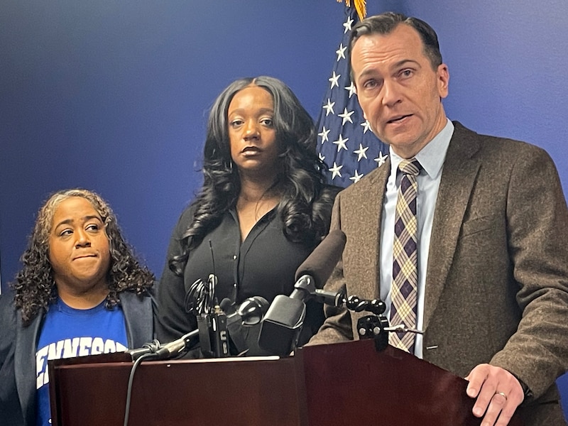 Three people stand in front of a blue wall and in front of a podium for a press conference.