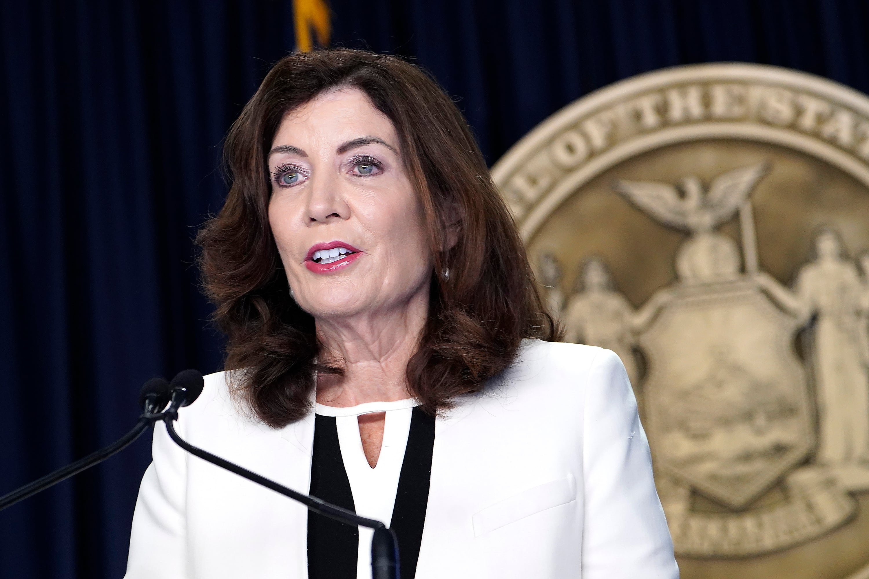 A woman with short brown hair and wearing a white suit speaks from a small microphone in front of a state seal.