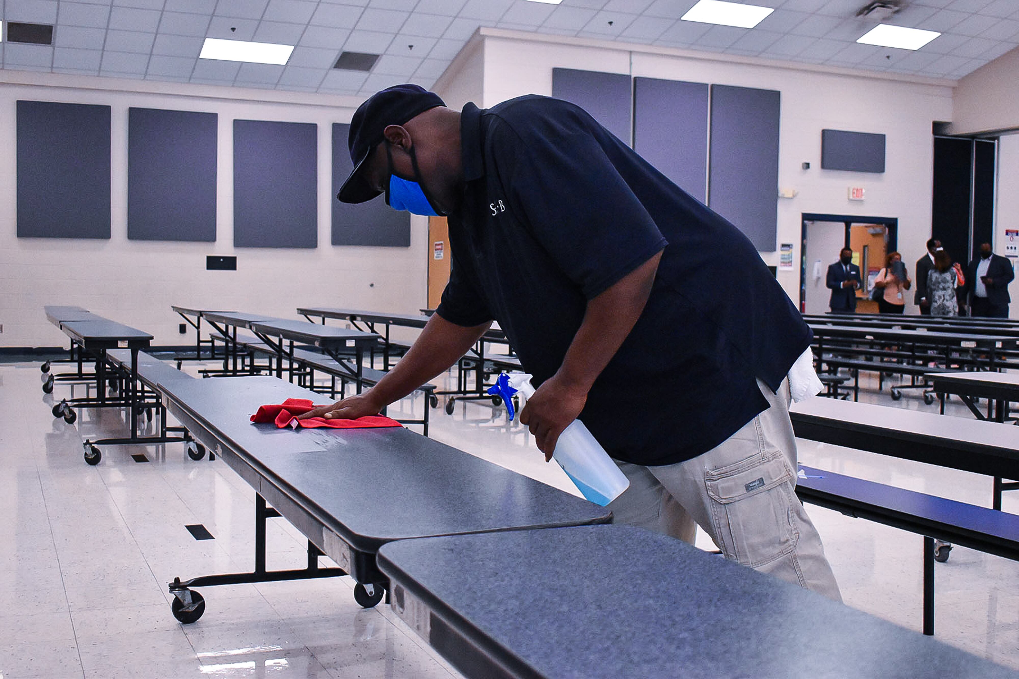 A Shelby County Schools employee sanitizes one of many long cafeteria tables while a group of people stand toward the back of the room.