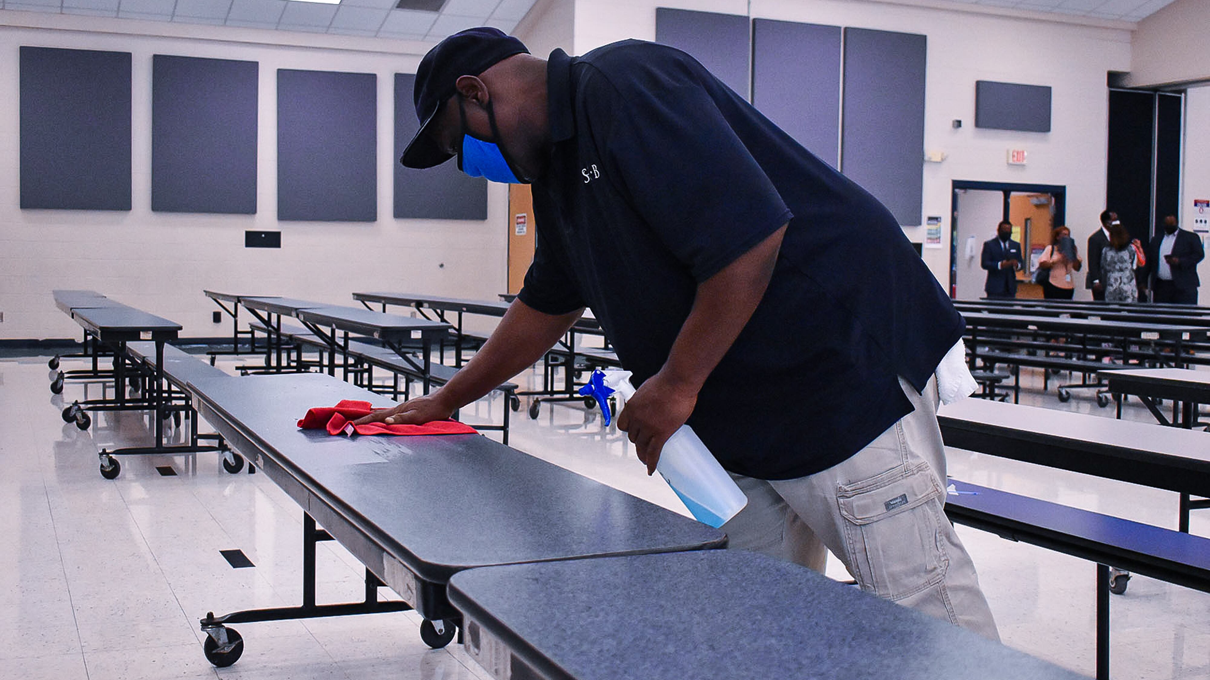A Shelby County Schools employee sanitizes one of many long cafeteria tables while a group of people stand toward the back of the room.
