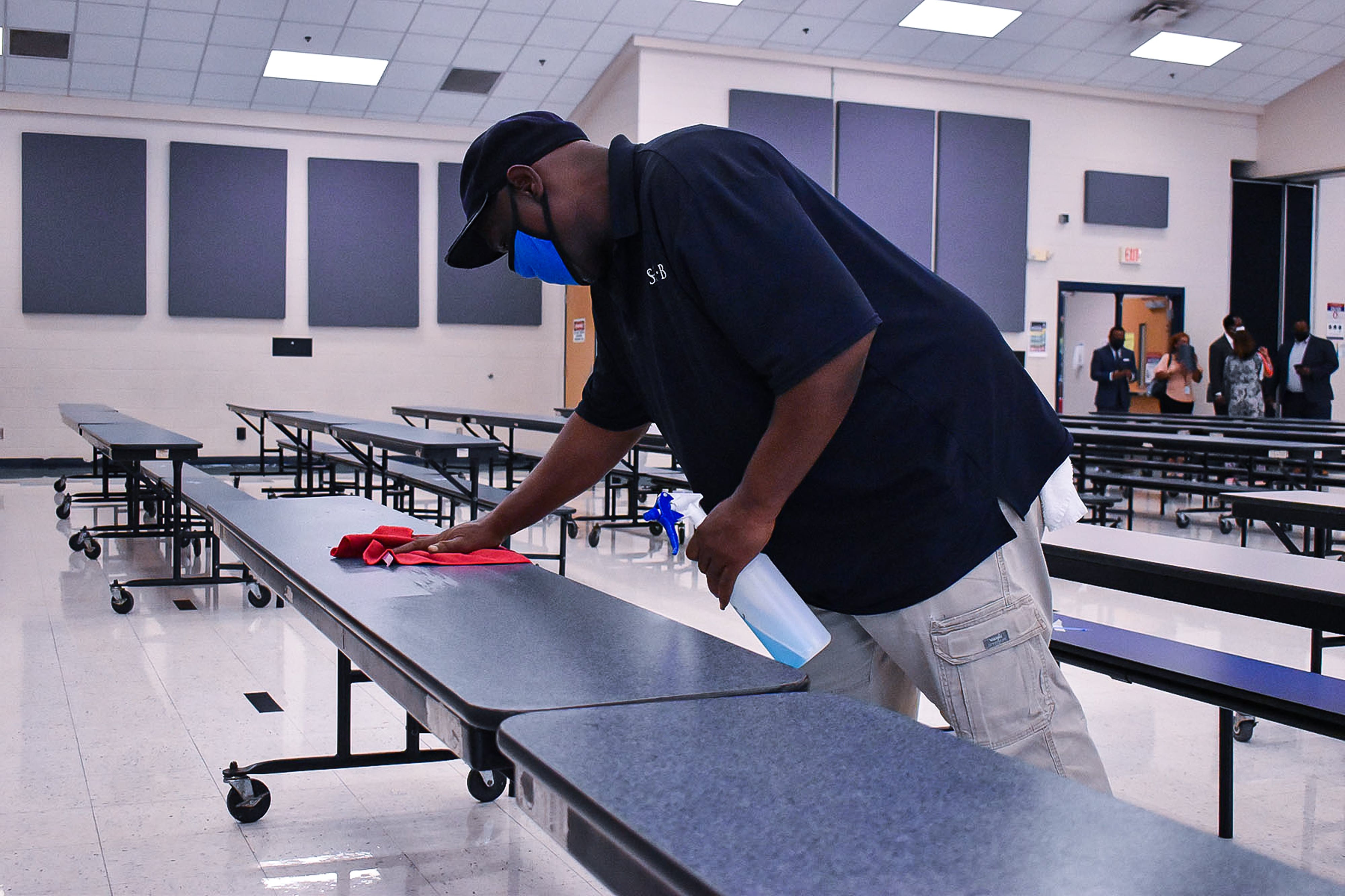 A Shelby County Schools employee sanitizes one of many long cafeteria tables while a group of people stand toward the back of the room.