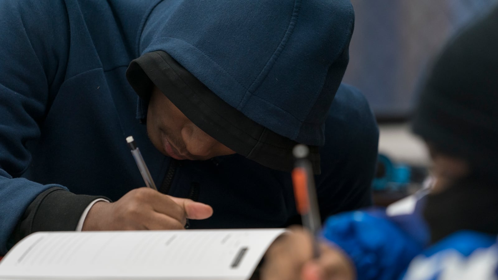 A student with a hood over their head writes into a instructional book.