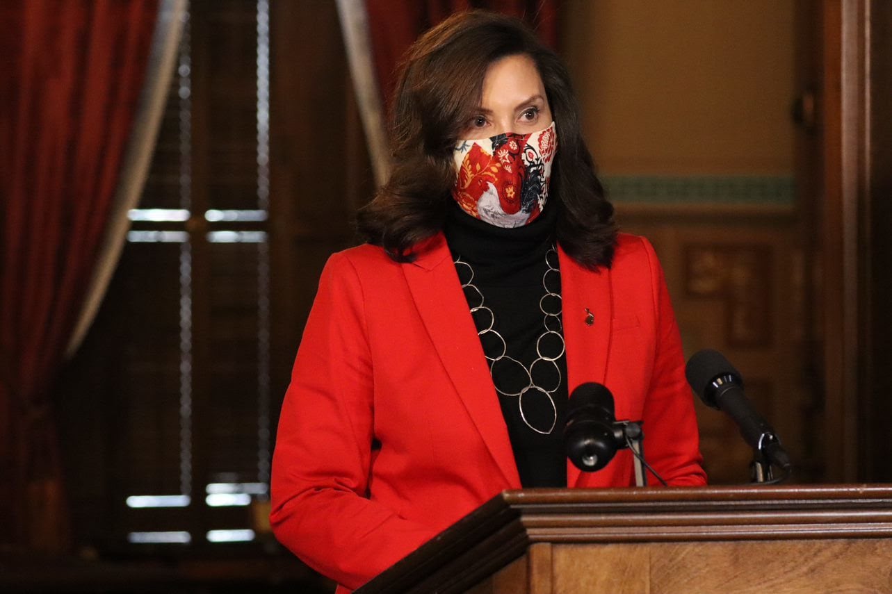 Michigan Gov. Gretchen Whitmer, wearing a mask and red jacket, stands at a podium