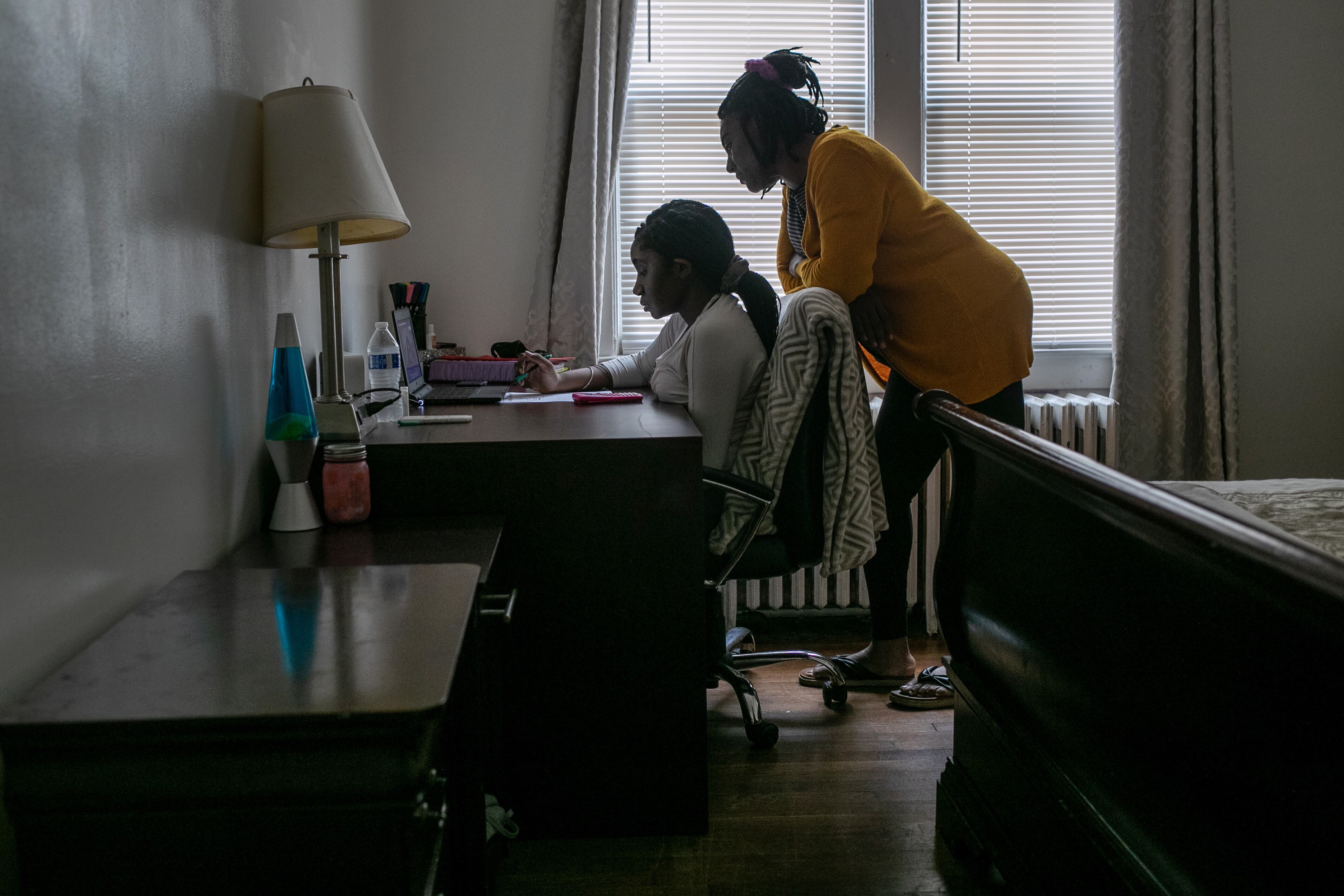 A mother leans over her daughter as she works in a bedroom, with the bed and a dresser in the foreground.