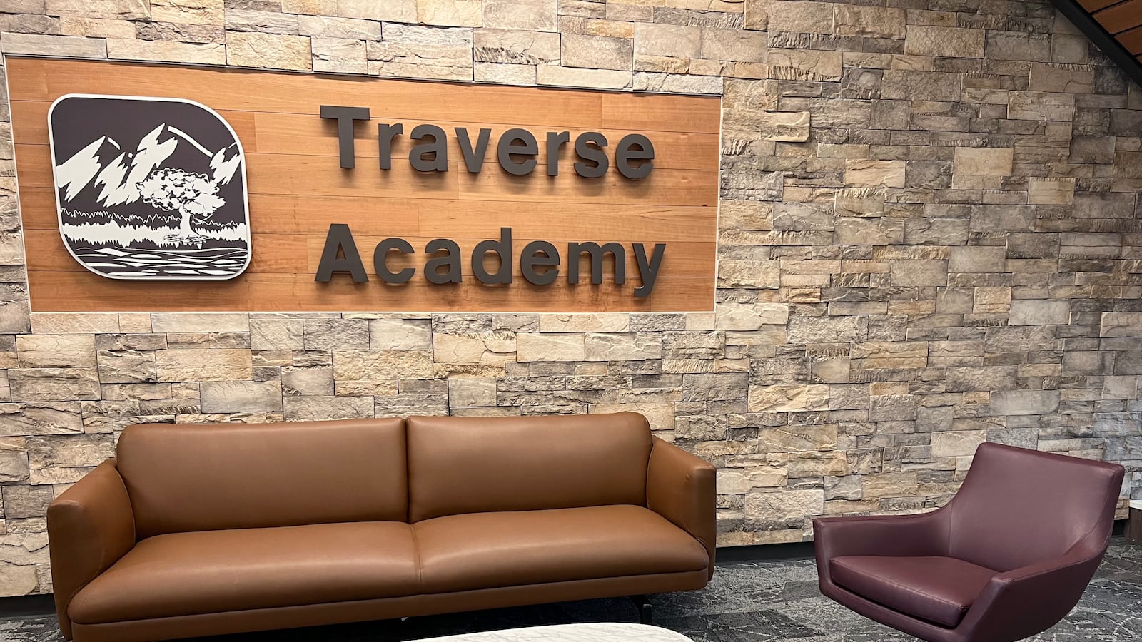 A brown couch and read chair sit in front of a wooden sign that hangs on the wall and reads “Traverse Academy.”