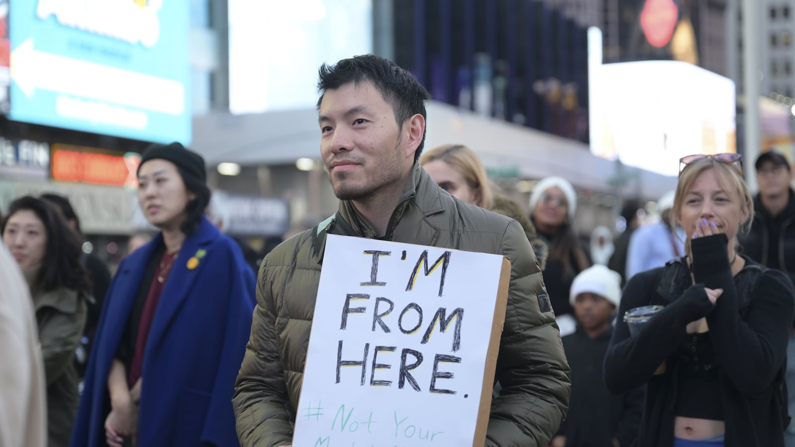 Asian Americans are gathered at the Times Square. A man holds a sign that says, “I’m From Here.”