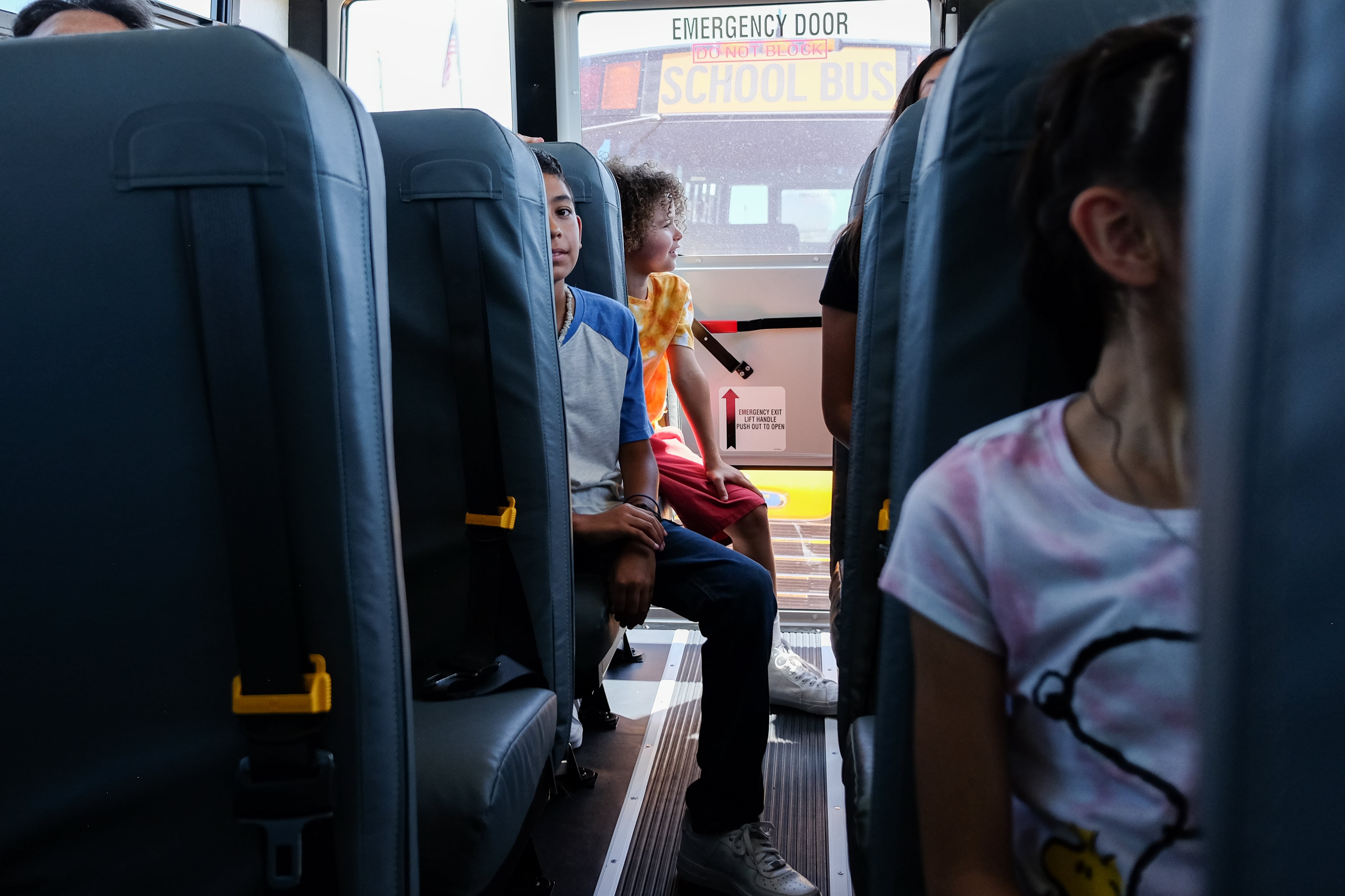A boy’s face can be seen peering out from a school bus seat. Other children are in the surrounding seats. The view is from inside the bus, looking down the center aisle. Another school bus can be seen out the back window.