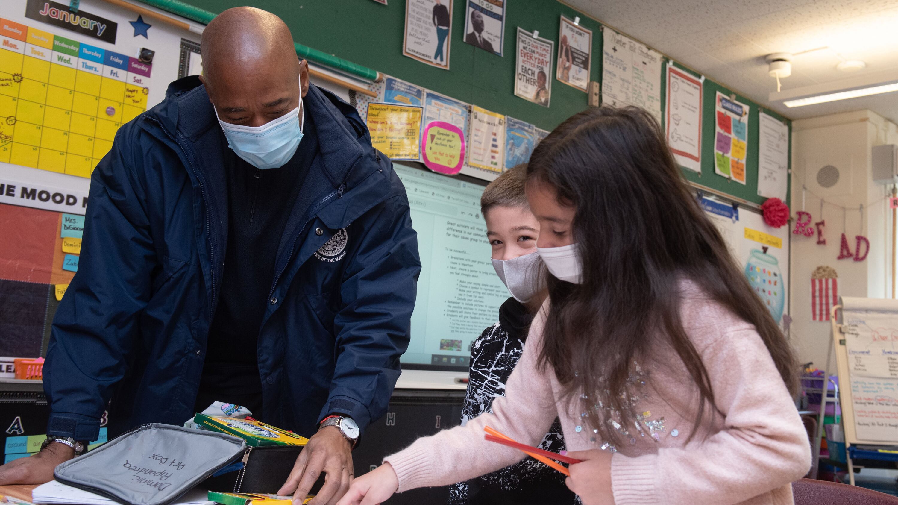 A man in a mask and a blue jacket looks at school work in a classroom with two children, also in masks. 