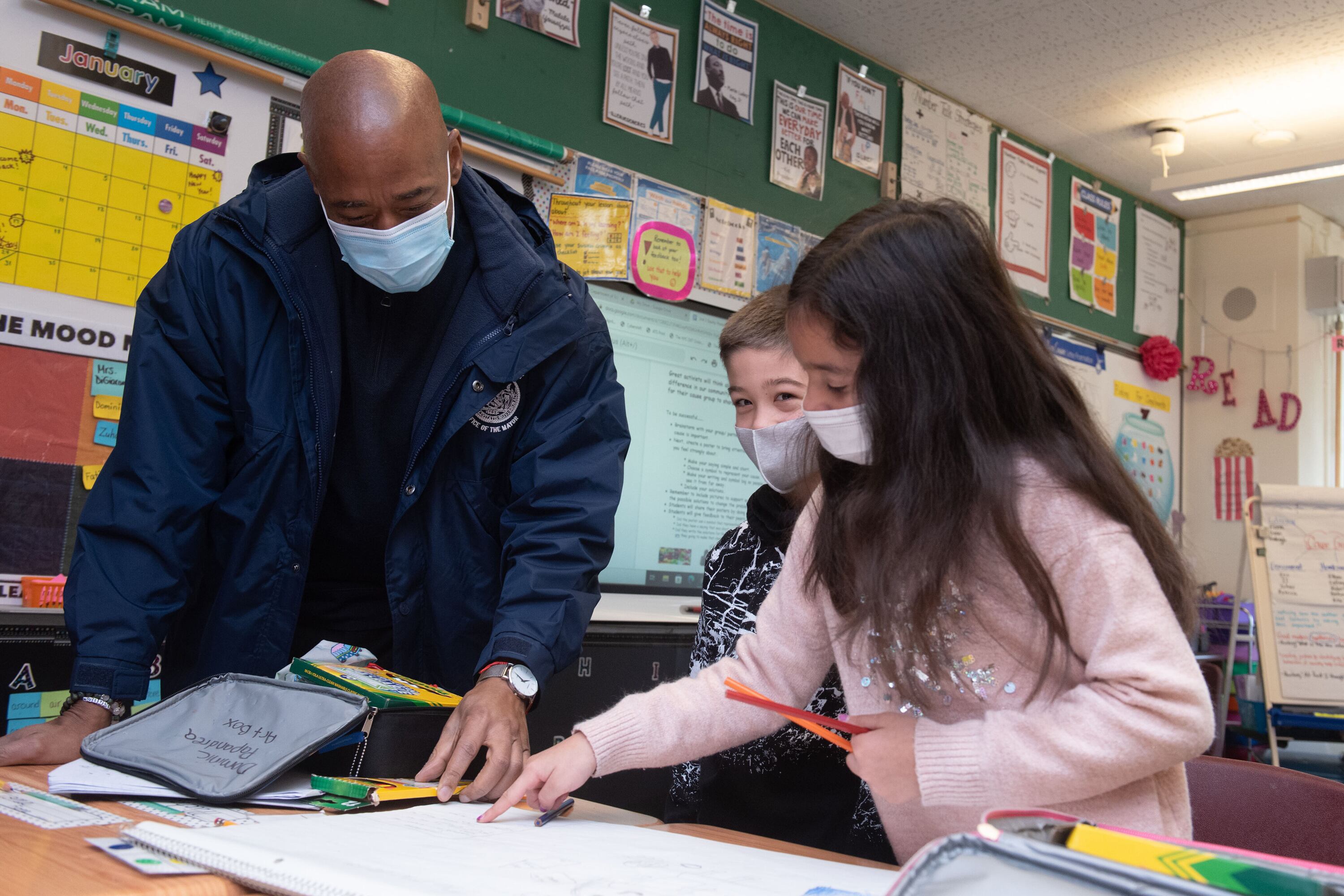 A man in a mask and a blue jacket looks at school work in a classroom with two children, also in masks.