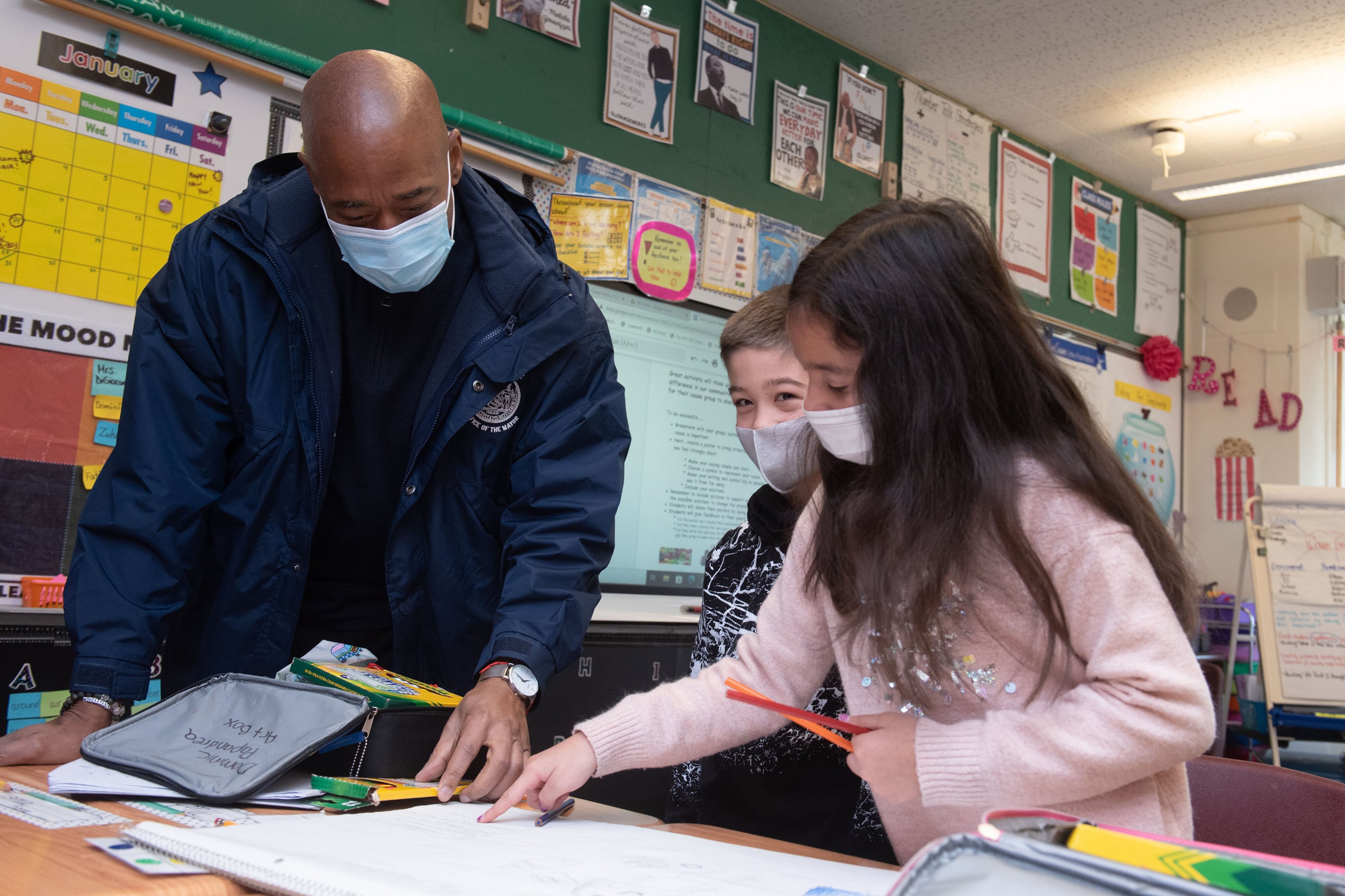 A man in a mask and a blue jacket looks at school work in a classroom with two children, also in masks. 