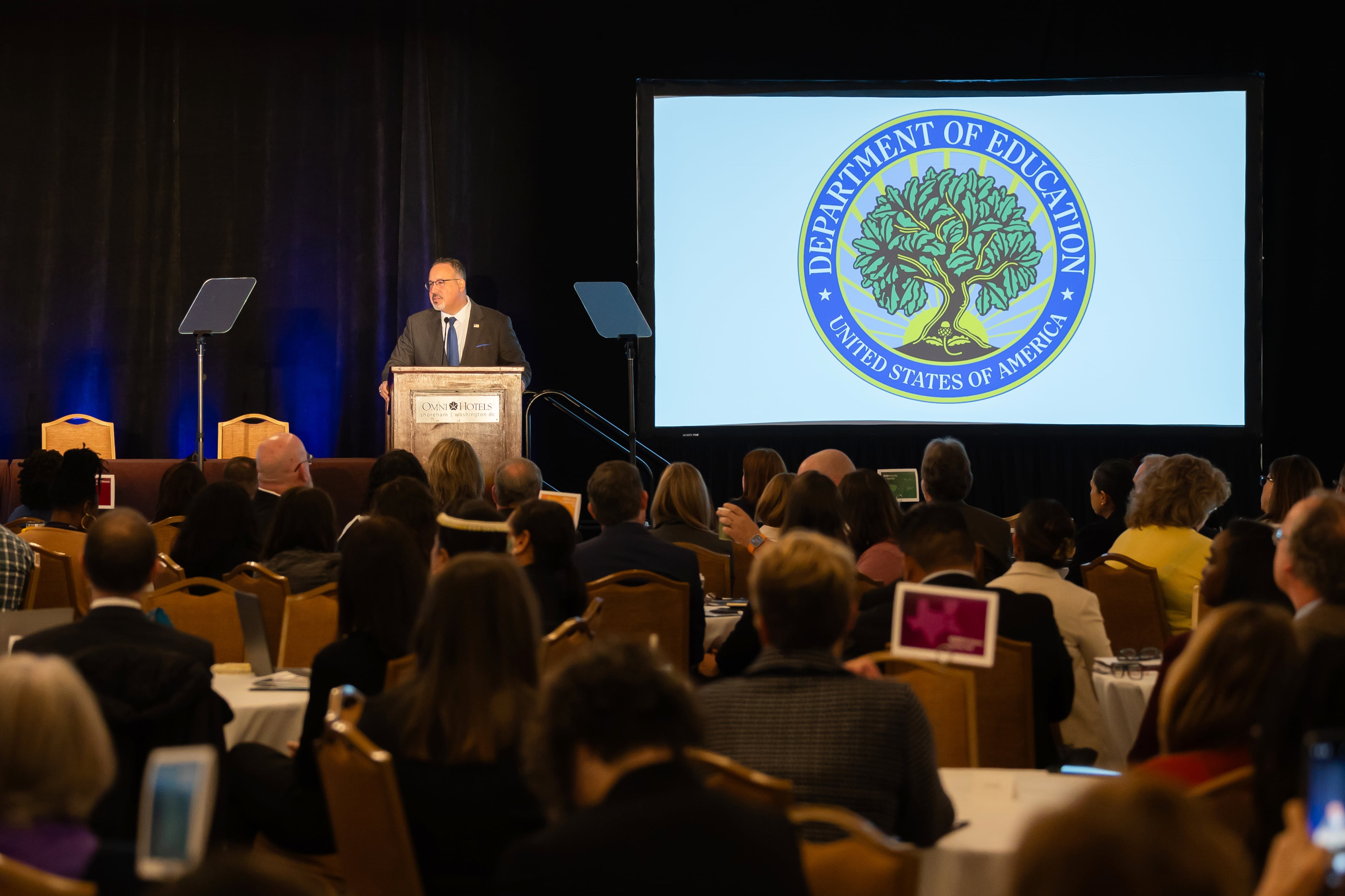 A man wearing a dark suit speaks from a wooden podium on a stage to an audience full of people. There is a large projection of the U.S. Department of Education's seal next to the speaker.