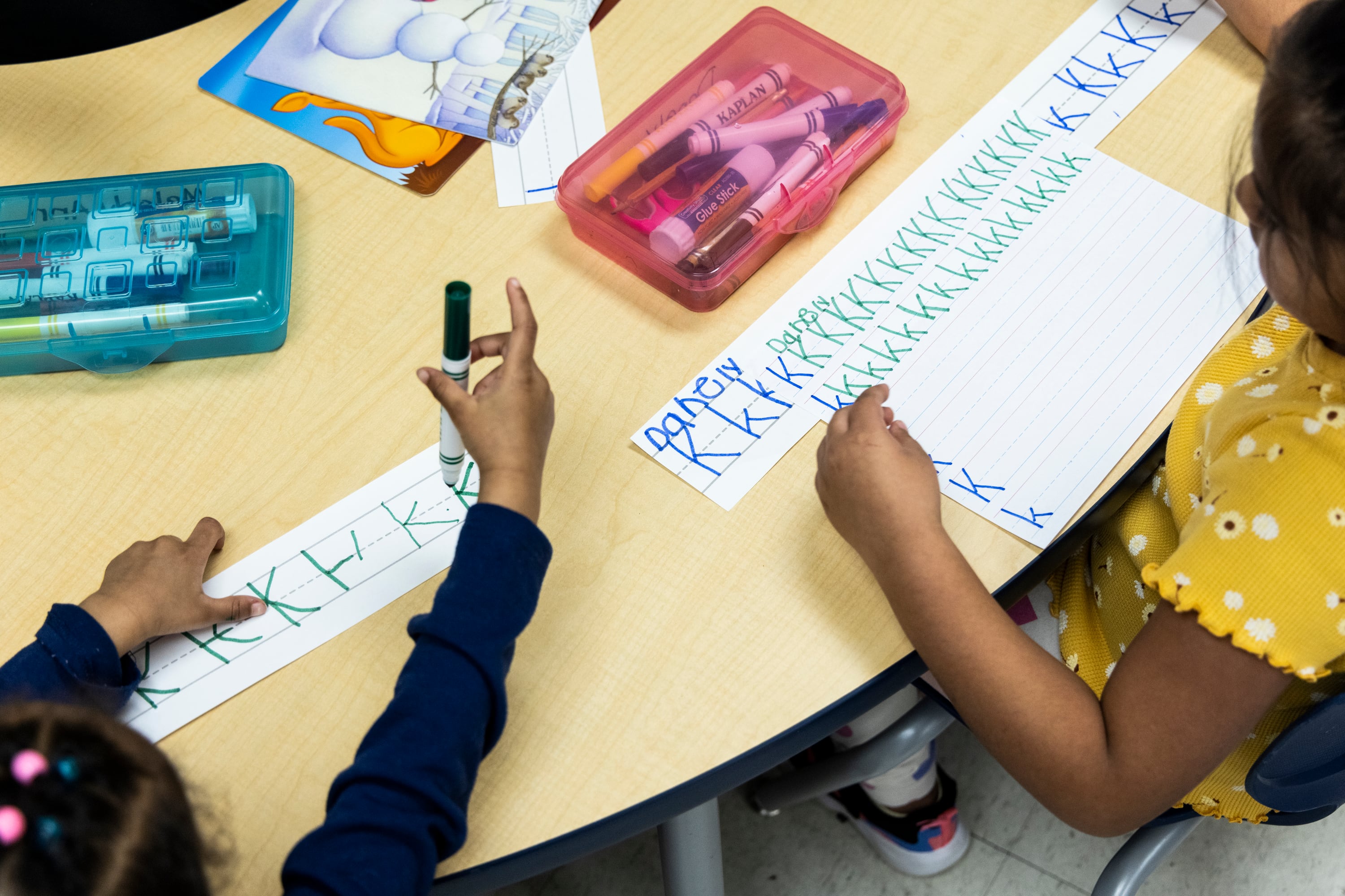 A bird's eye view of two young students working at a table writing their alphabet.